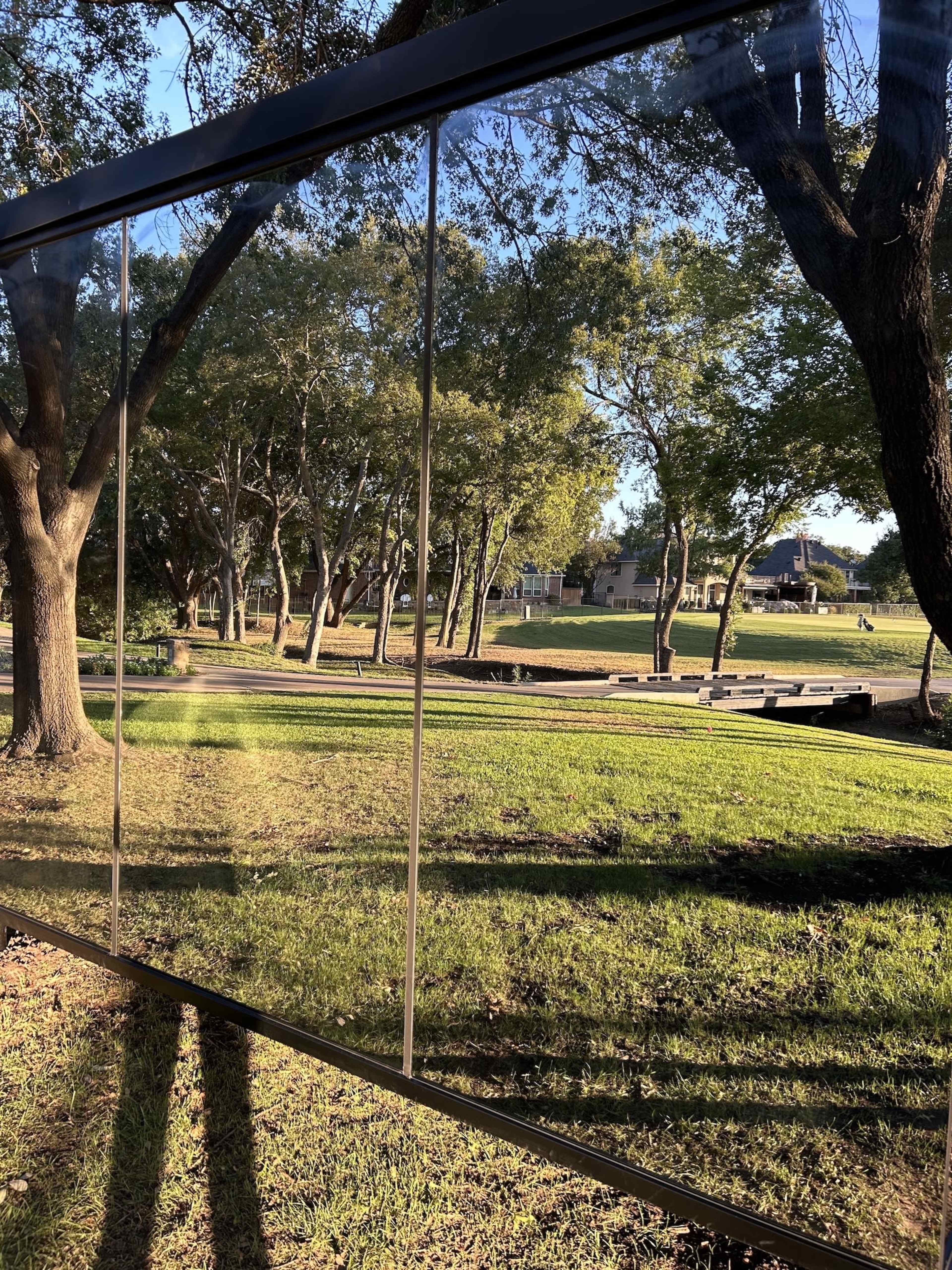 A large glass panel reflects a park scene with trees and a pathway lined with benches.
