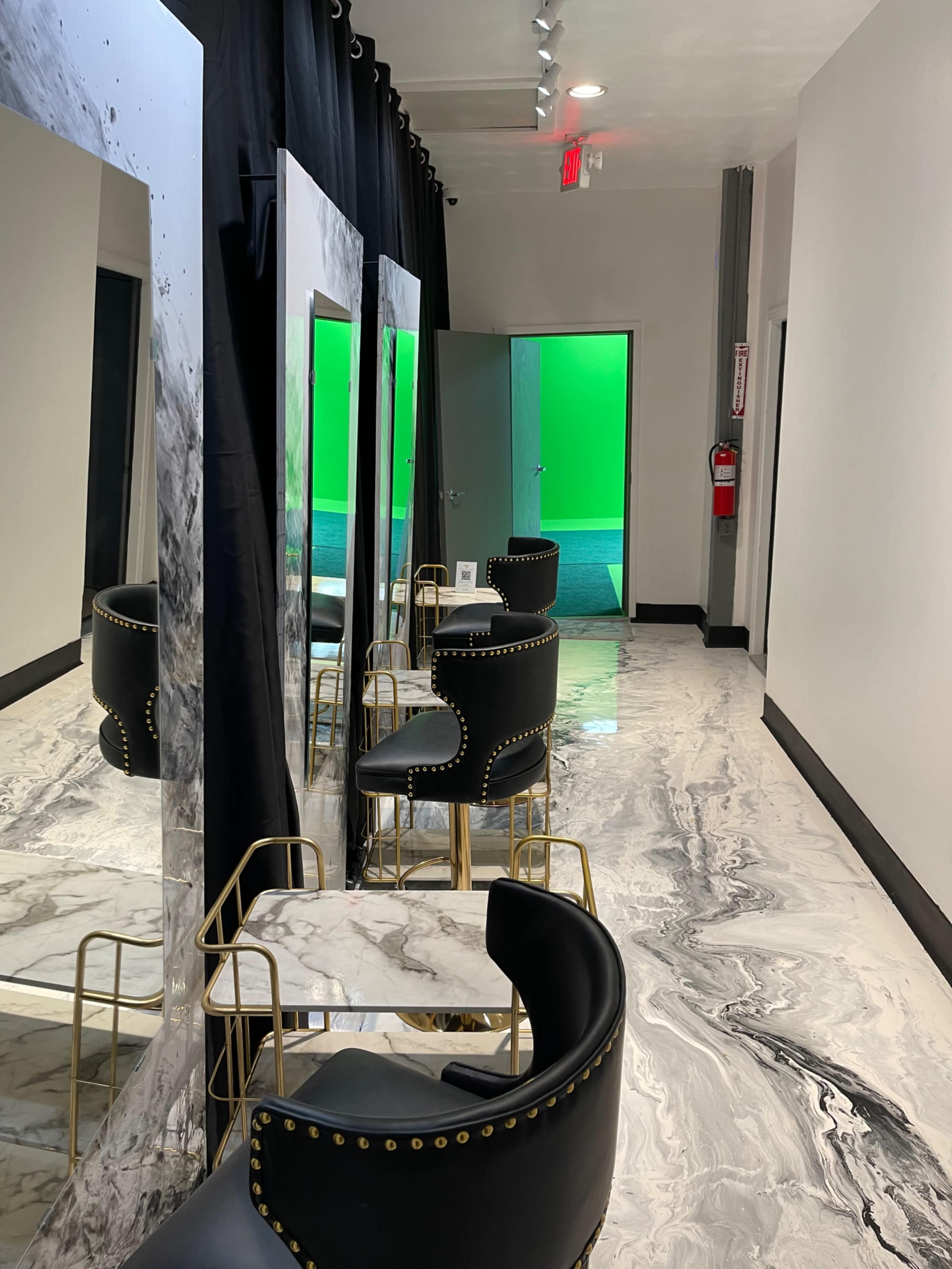 A salon interior featuring black and gold chairs arranged around a marble table, with large mirrors and a green backdrop visible through an open door.