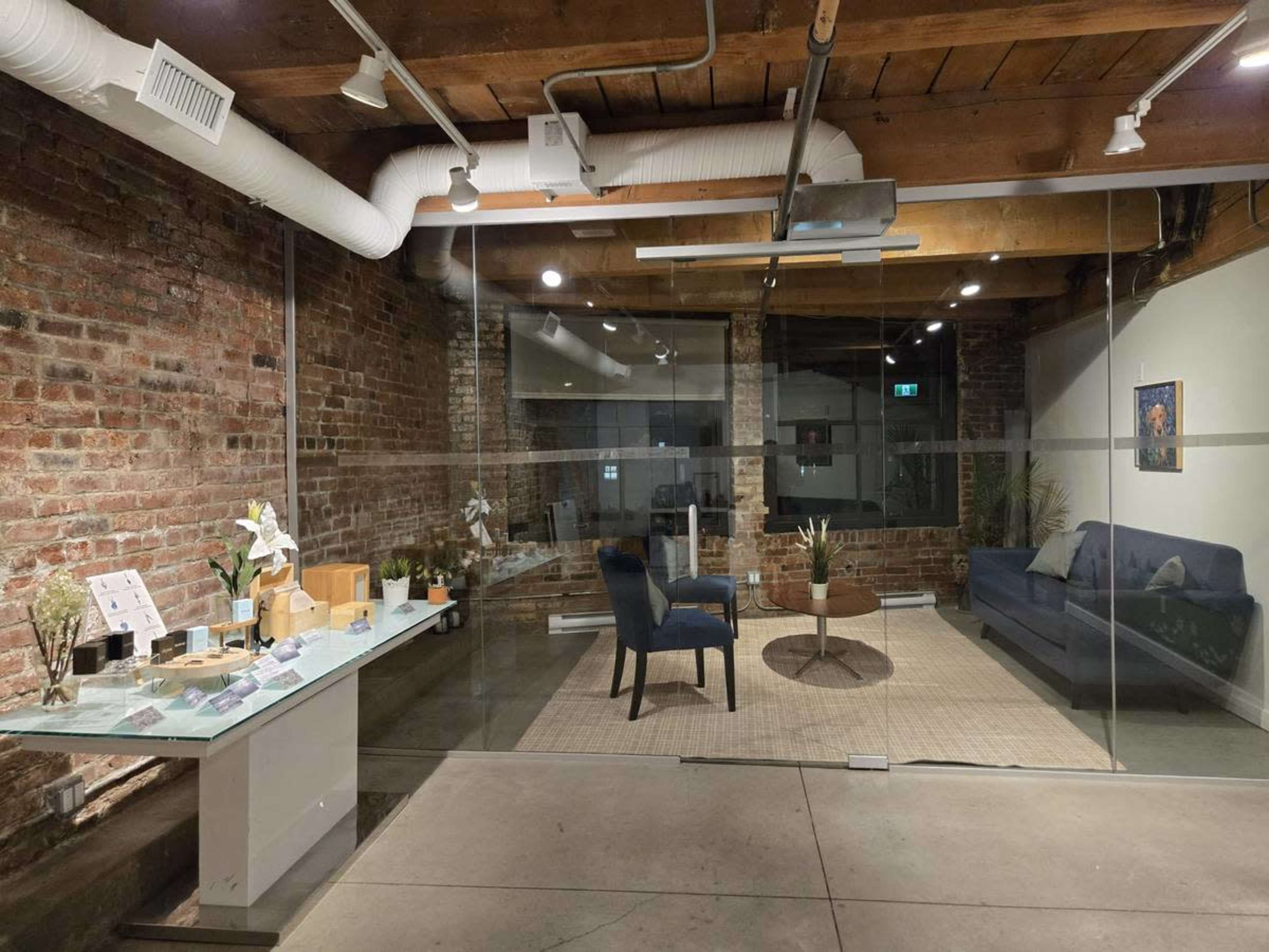 The image shows a modern interior space with exposed brick walls, a glass partition, and a seating area featuring a blue couch, circular coffee table, and a decorative rug.