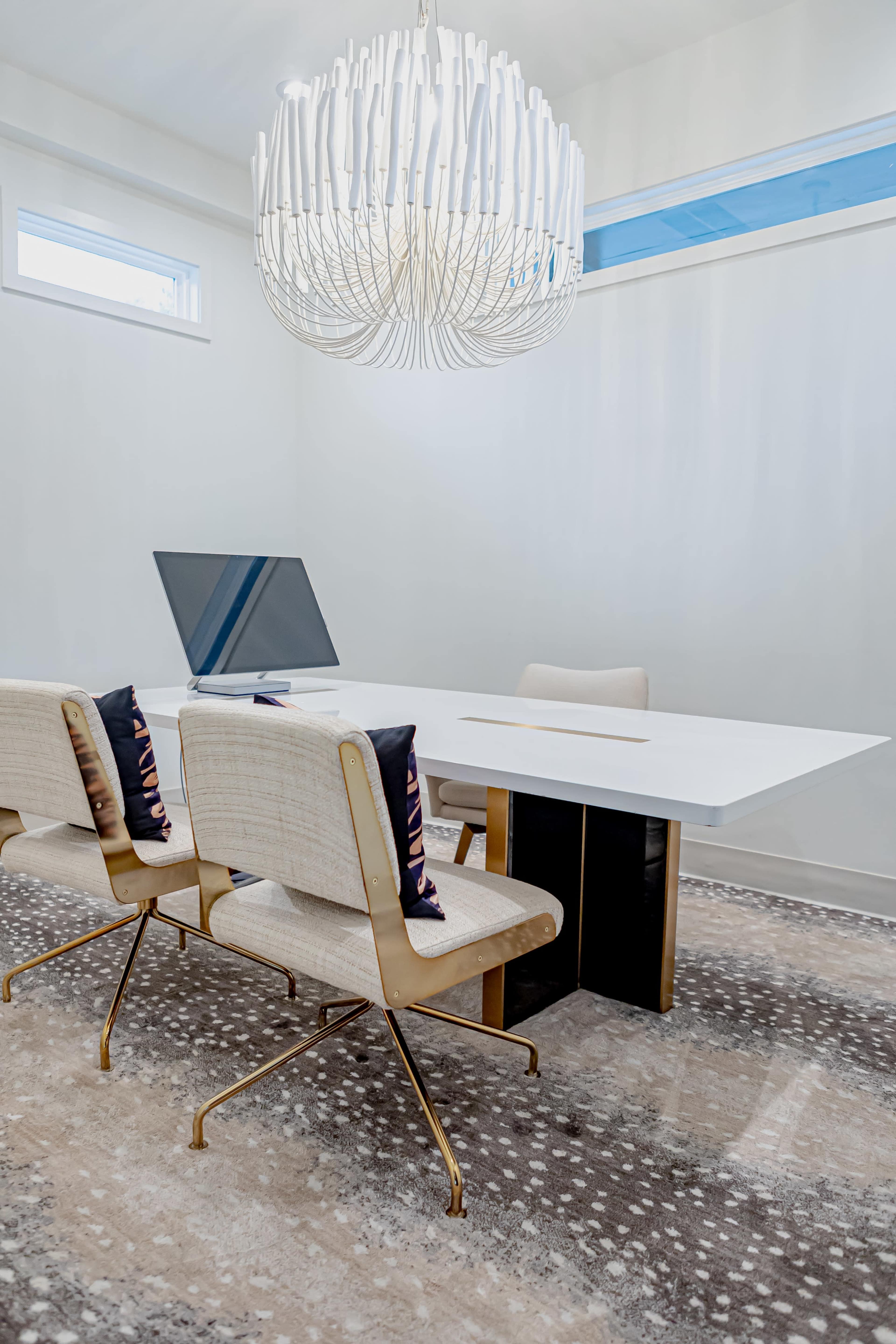 A modern office space features a sleek white table, three beige chairs with cushions, and a large chandelier above, all situated on a patterned rug.