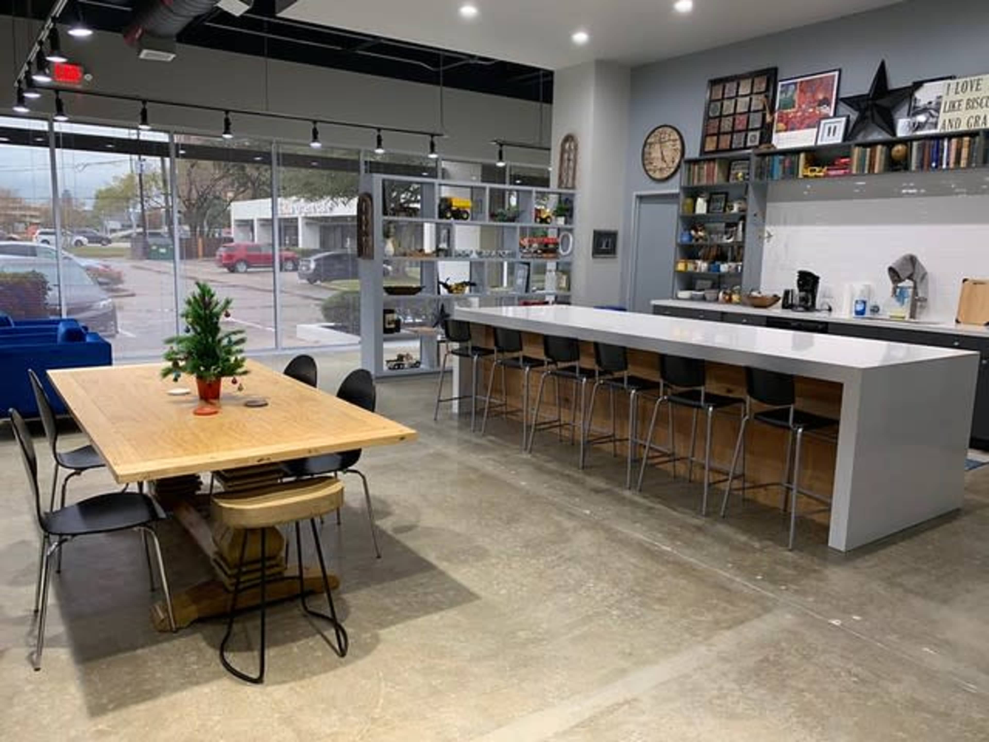 The image shows a modern kitchen and dining area featuring a long table and a bar with high stools, set in a spacious, well-lit environment.
