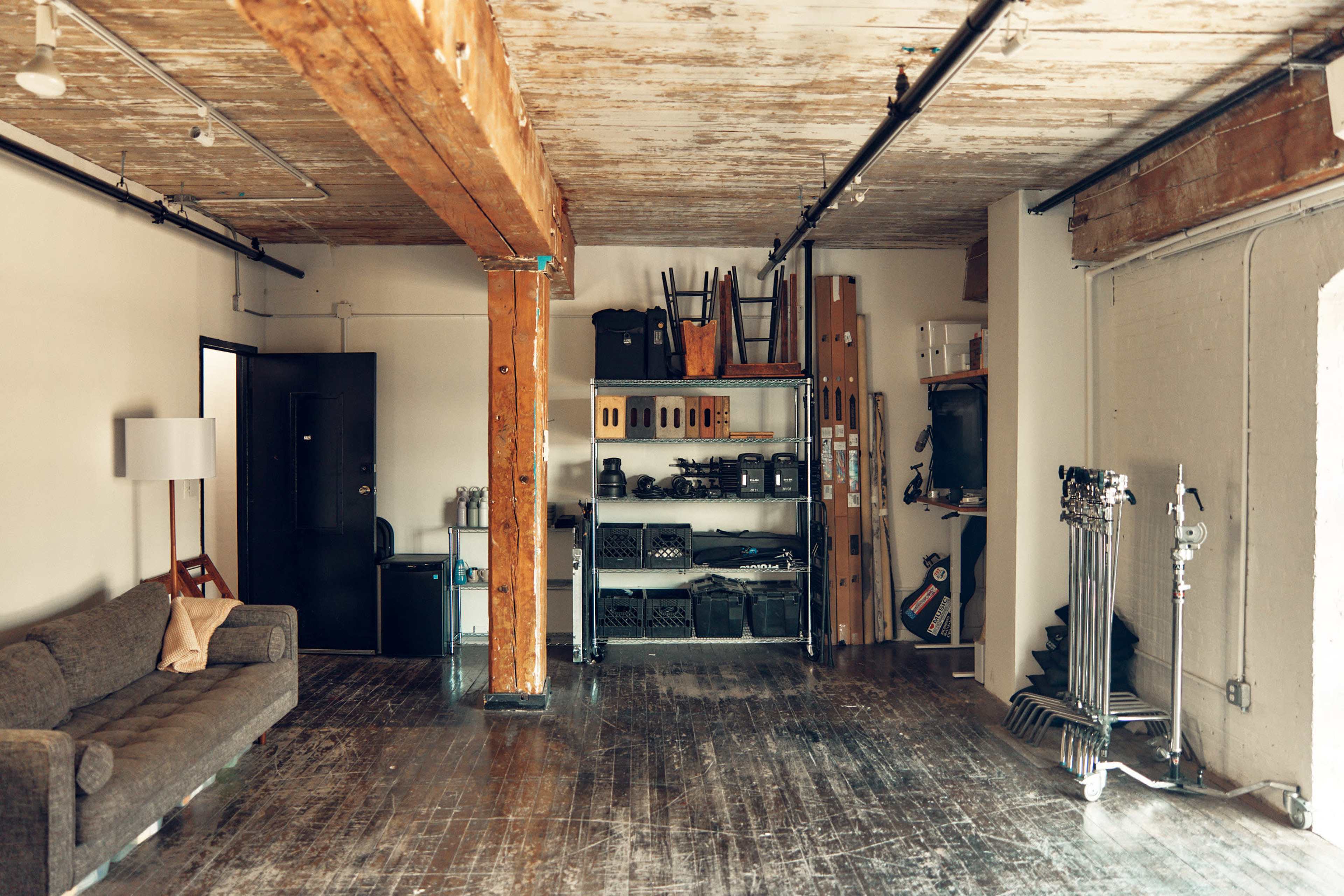 The image shows a spacious room with exposed wooden beams, a grey sofa, shelving units filled with equipment, and a doorway leading to another space.