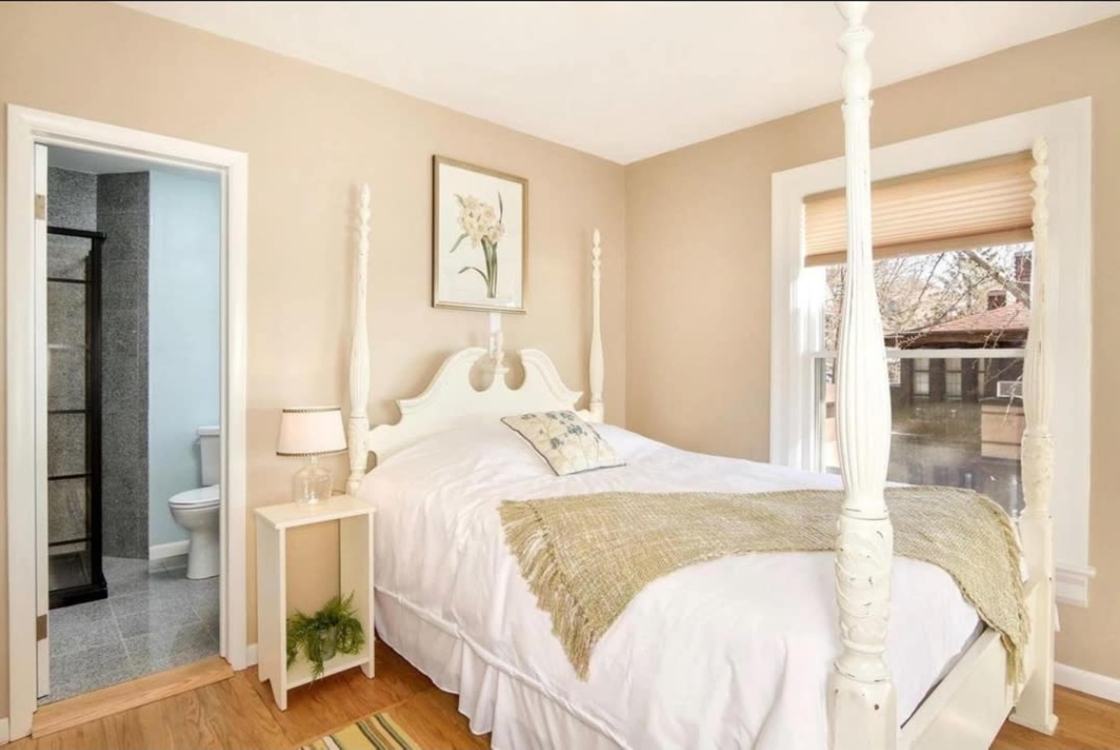 A bedroom with a four-poster bed, a side table with a lamp, and a bathroom entrance in the background.