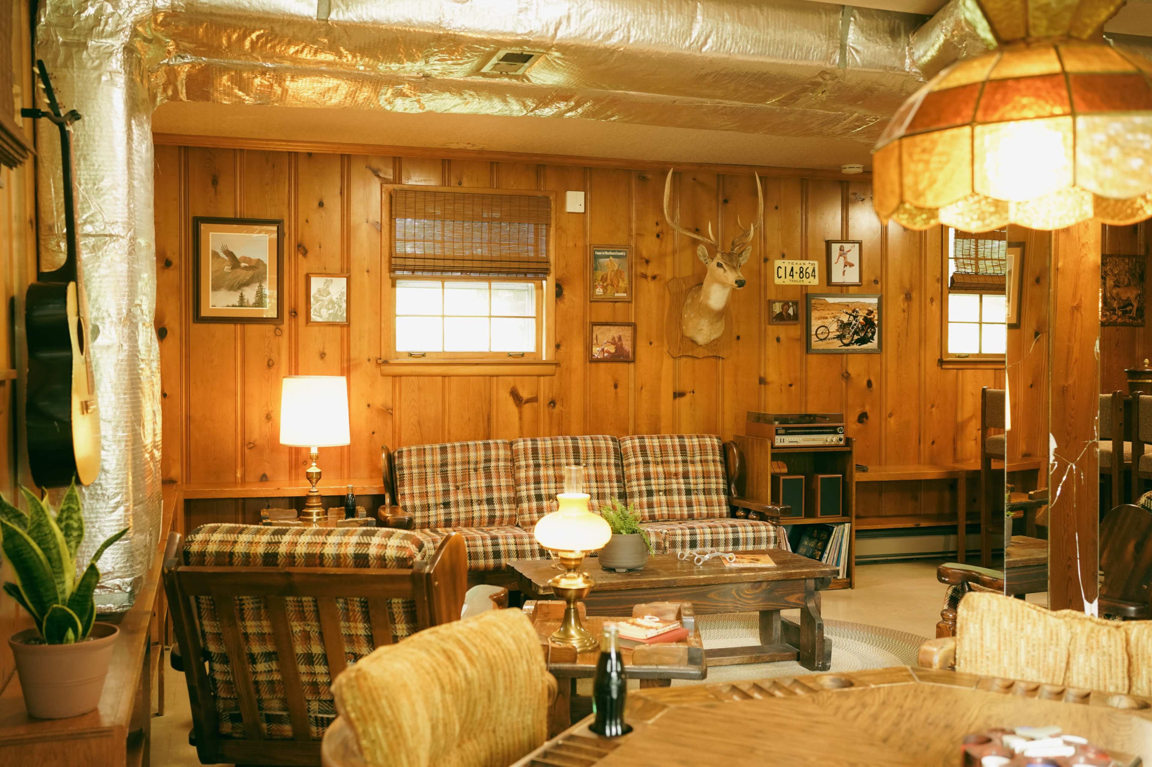 The image shows a cozy wooden-paneled living room with a plaid sofa, a wooden dining table, a mounted deer head on the wall, and various decorative items.