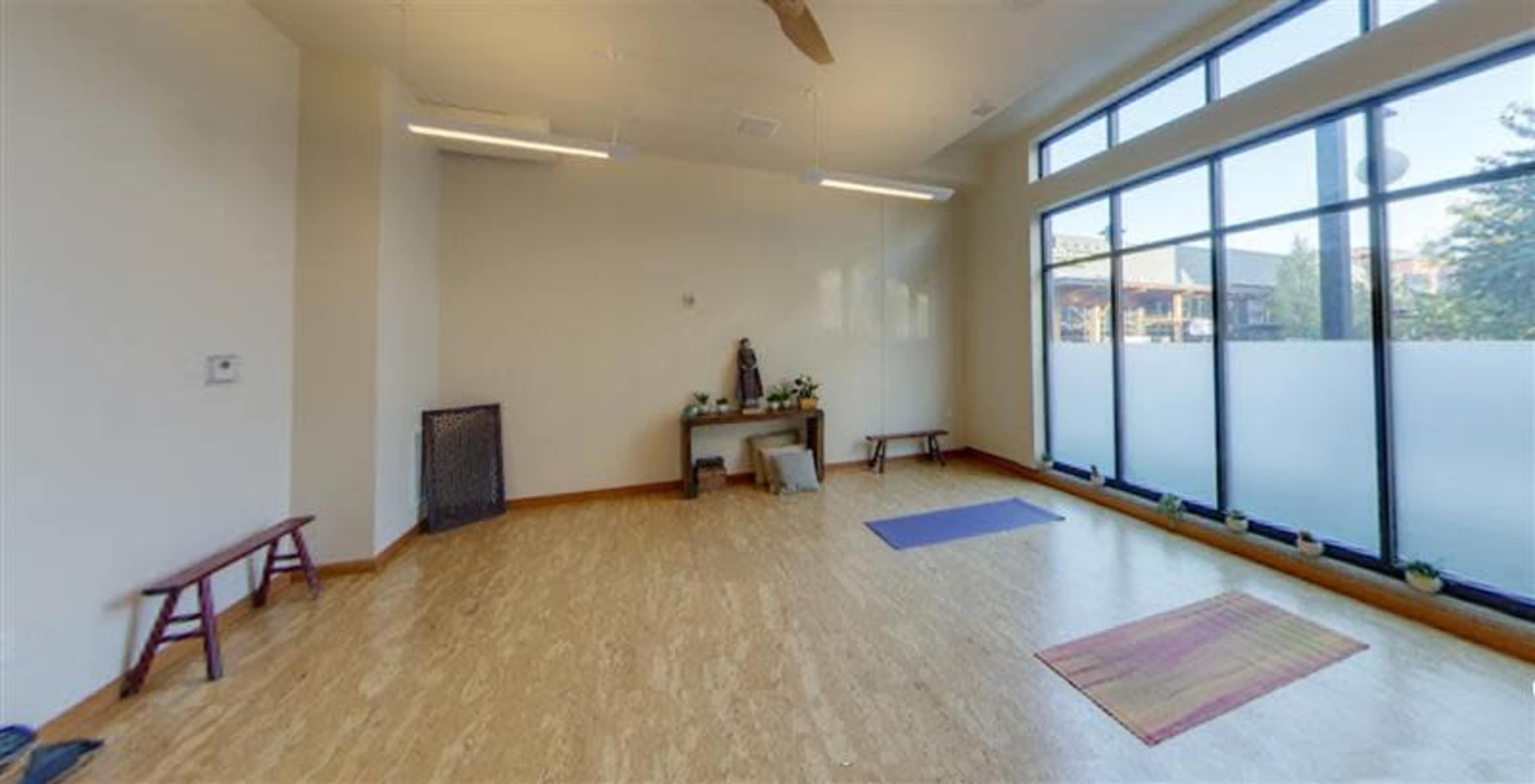 The image shows a spacious, empty studio with wooden flooring, two benches, and yoga mats placed on the ground, illuminated by natural light through large windows.