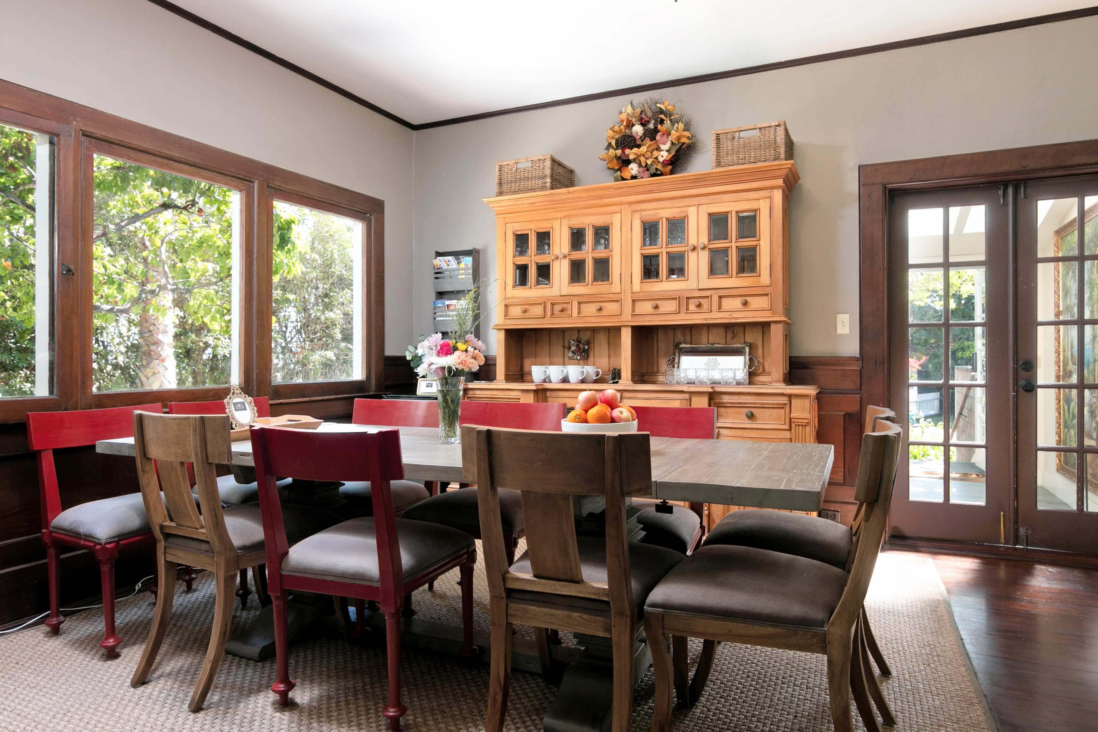 A wooden dining table surrounded by red and brown chairs is positioned in a well-lit dining room with large windows and a sideboard.