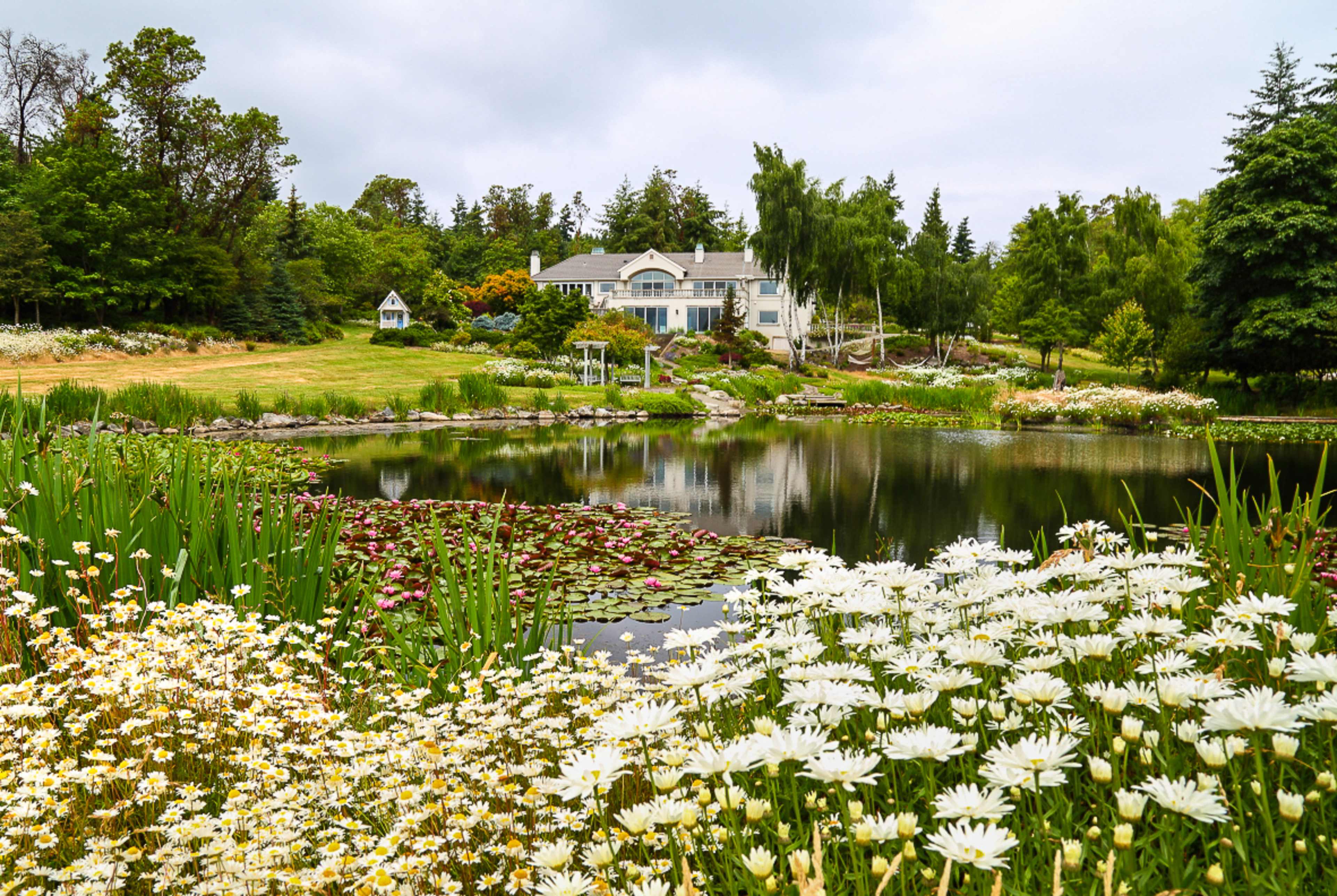 A large house is situated by a pond surrounded by blooming flowers and greenery.
