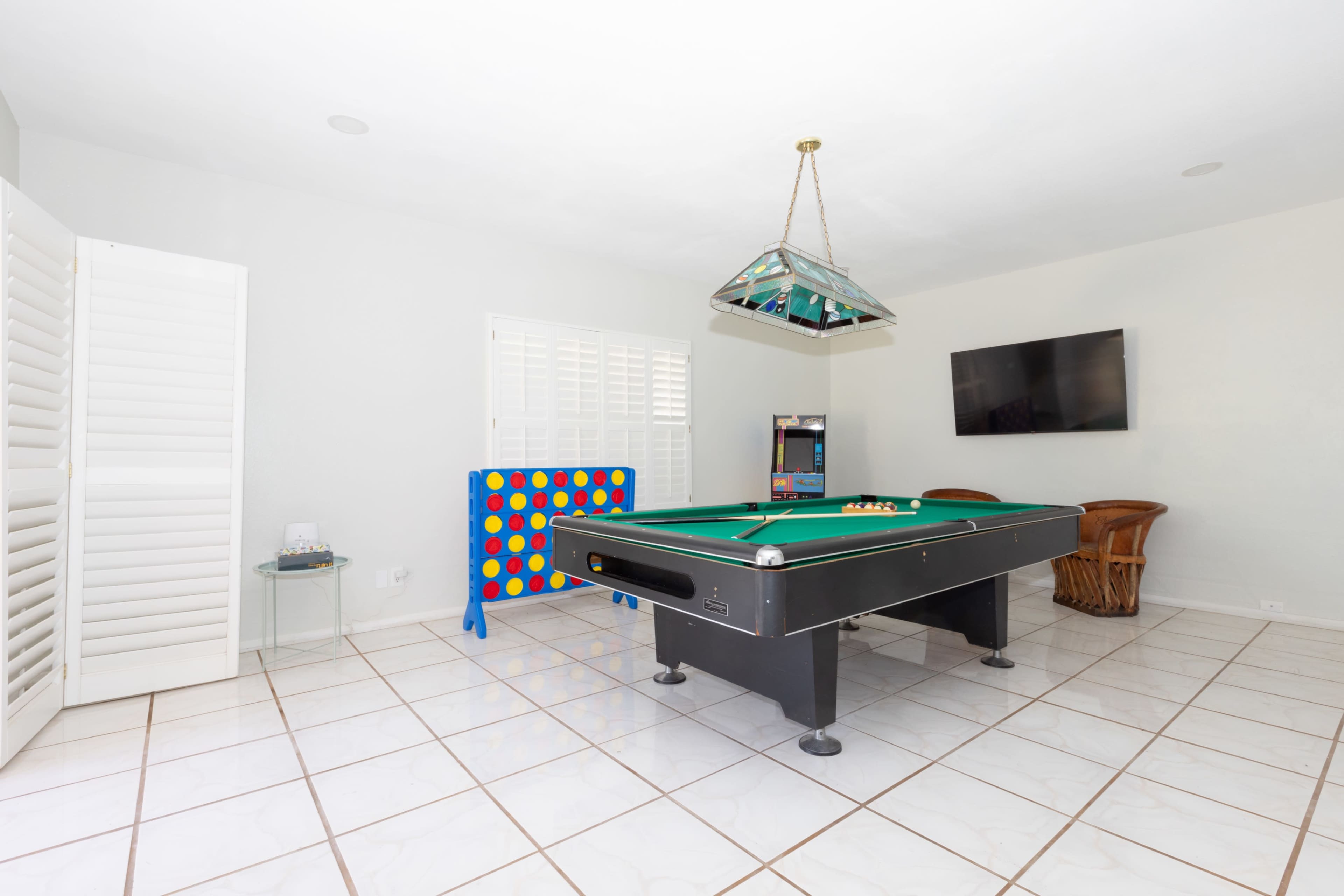 A game room featuring a pool table, colorful connect four game, a television mounted on the wall, and wicker chairs on a tiled floor.