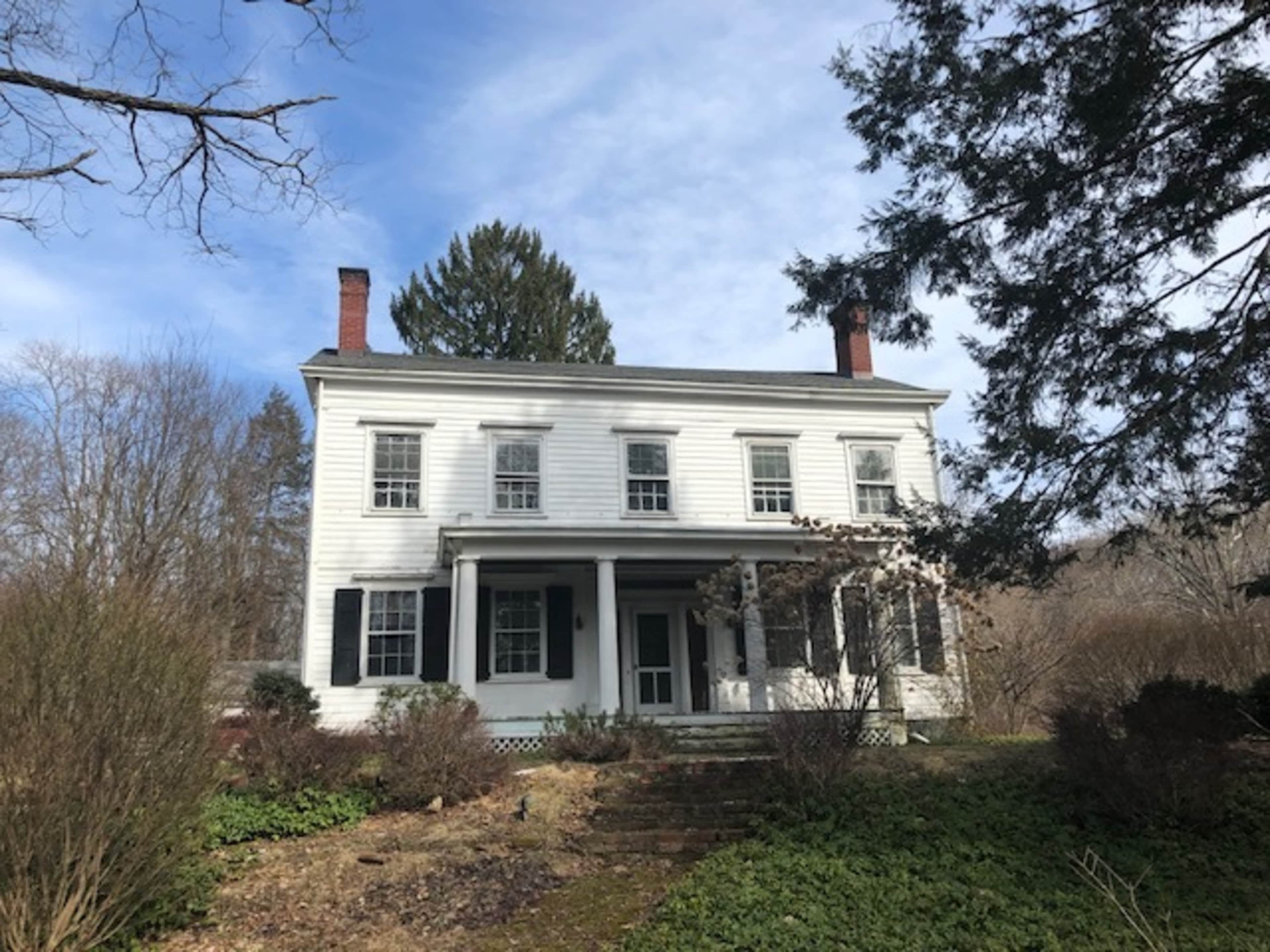 A two-story white colonial house with black shutters and a front porch is surrounded by overgrown shrubs and trees.