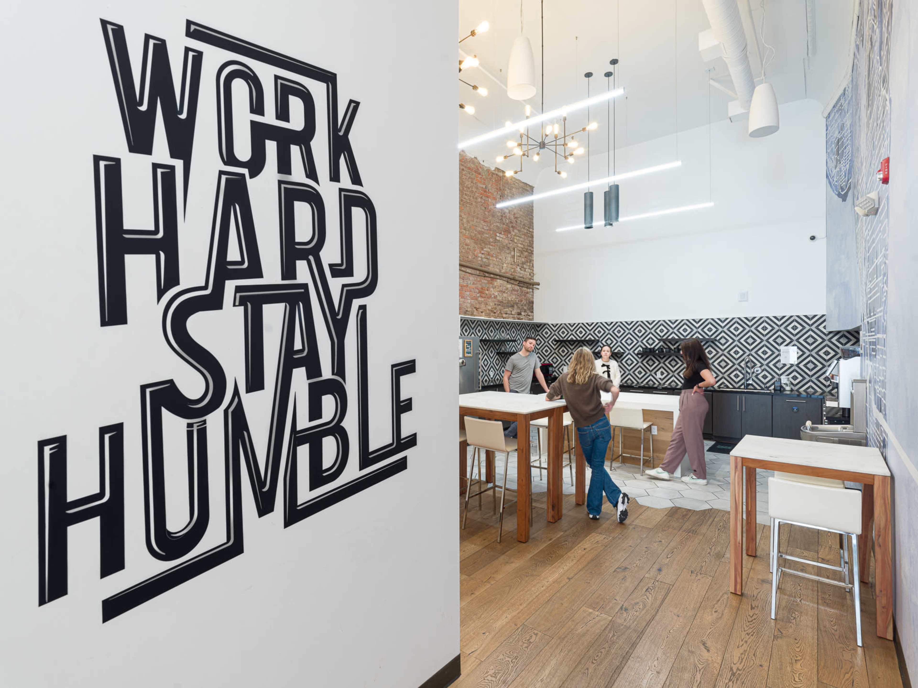 The image shows a modern office space with a motivational wall mural that says "WORK HARD STAY HUMBLE," and people seated around a large table in the background.