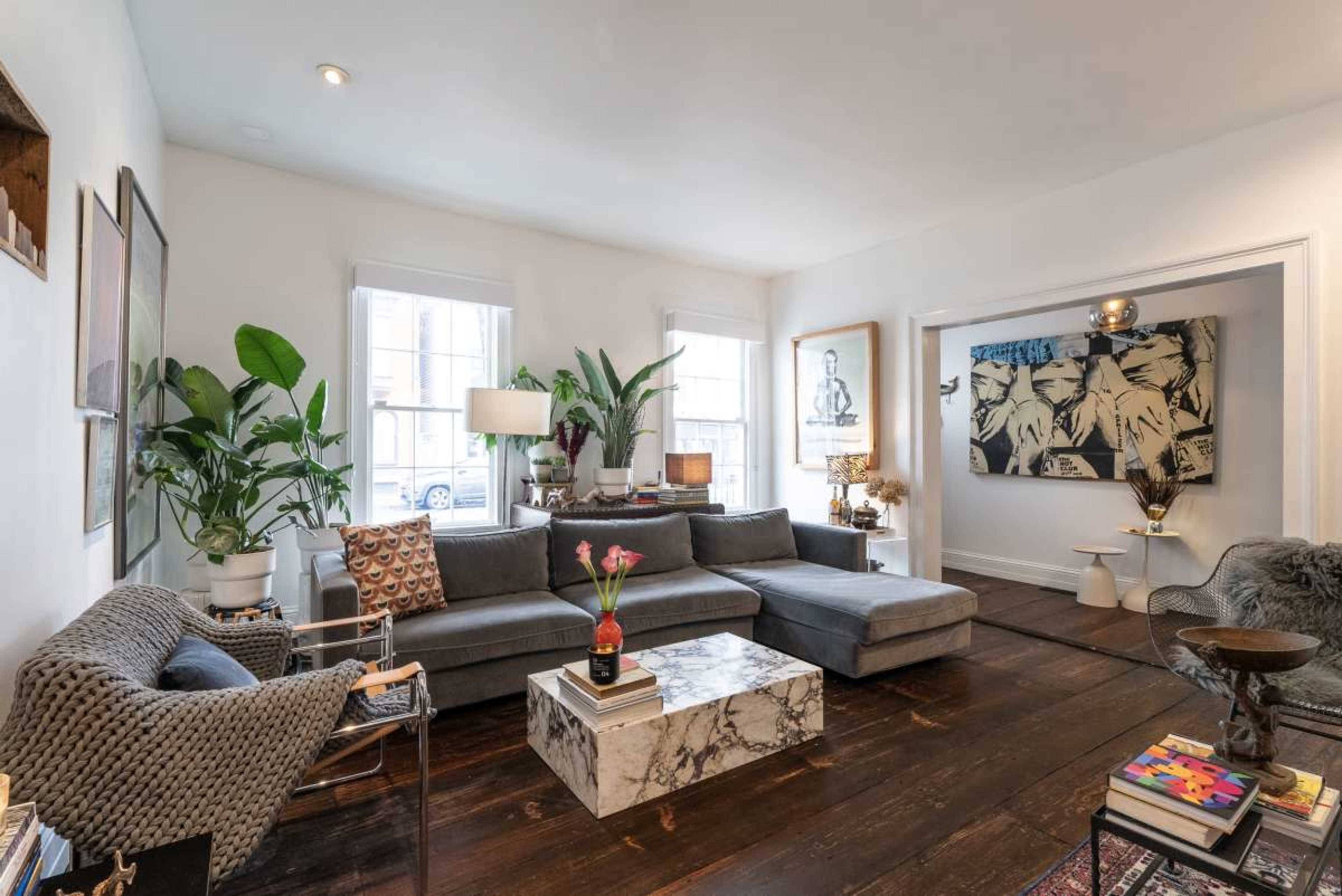 The living room features a sectional gray sofa, a marble coffee table, potted plants, and artwork on the walls, along with windows allowing natural light to enter.