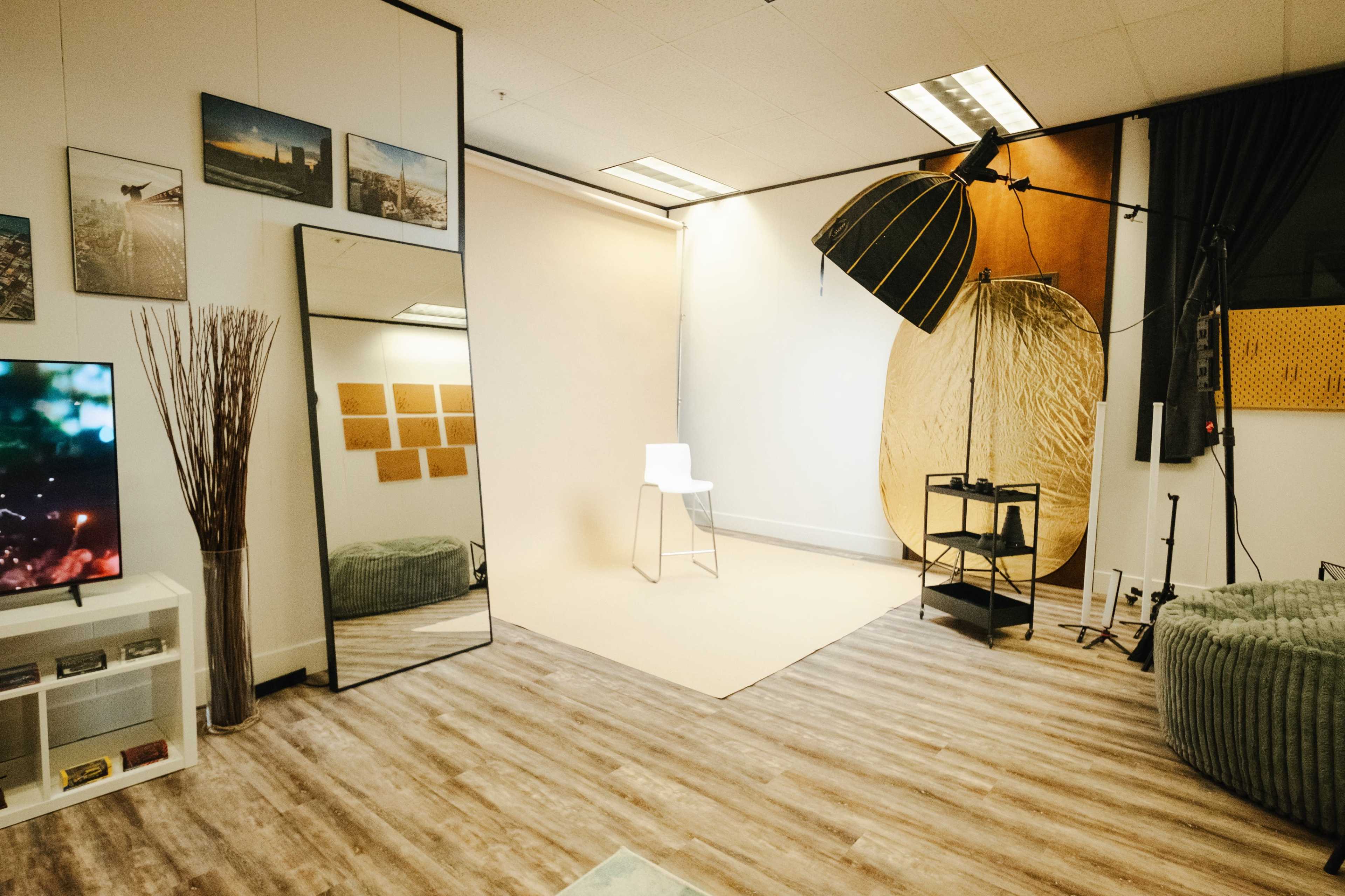 A photography studio equipped with a large mirror, a backdrop, a chair, a stand with lighting equipment, and a textured wall.