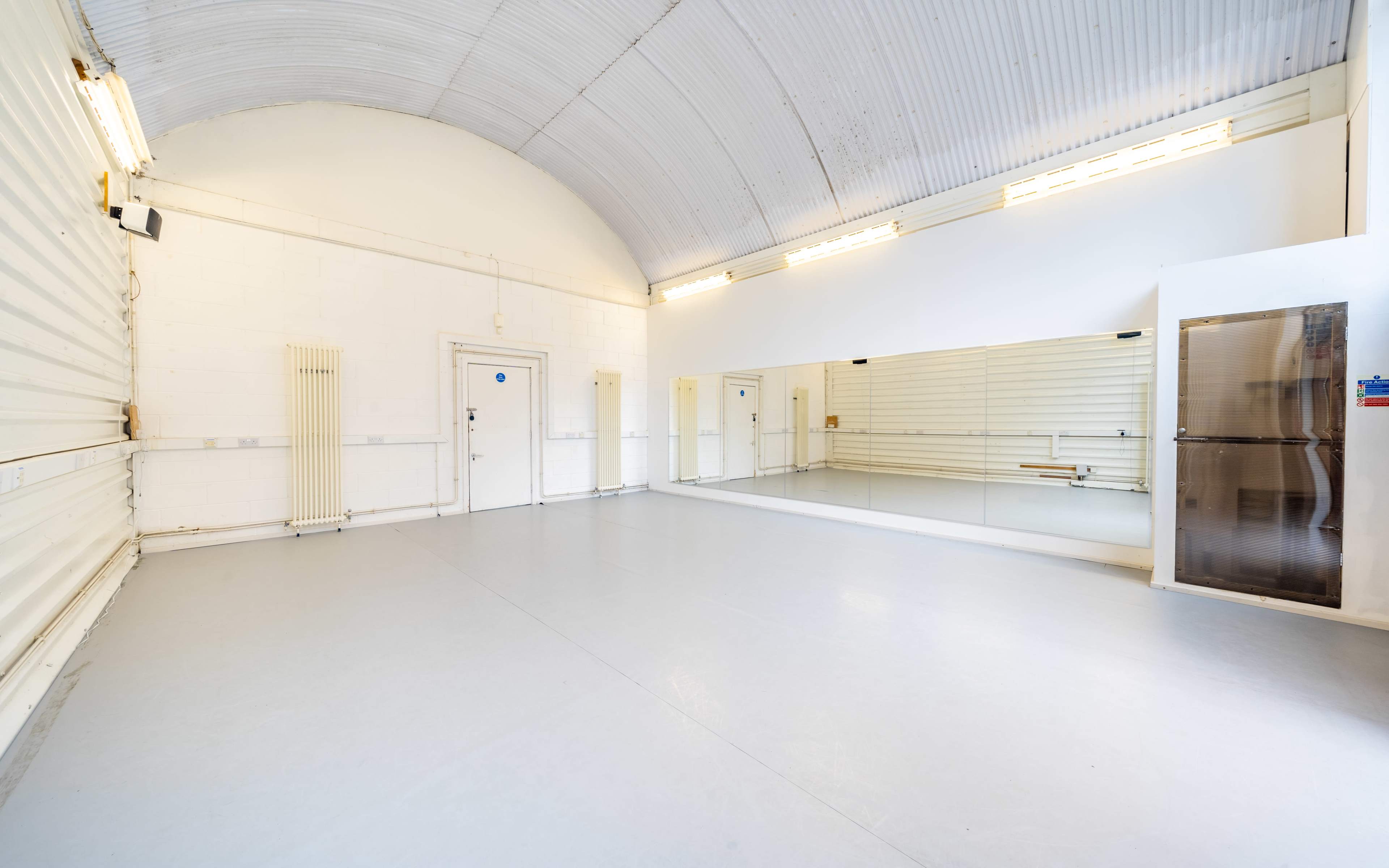 A spacious, empty room with a mirrored wall and white walls, featuring a high arched ceiling and a gray floor.