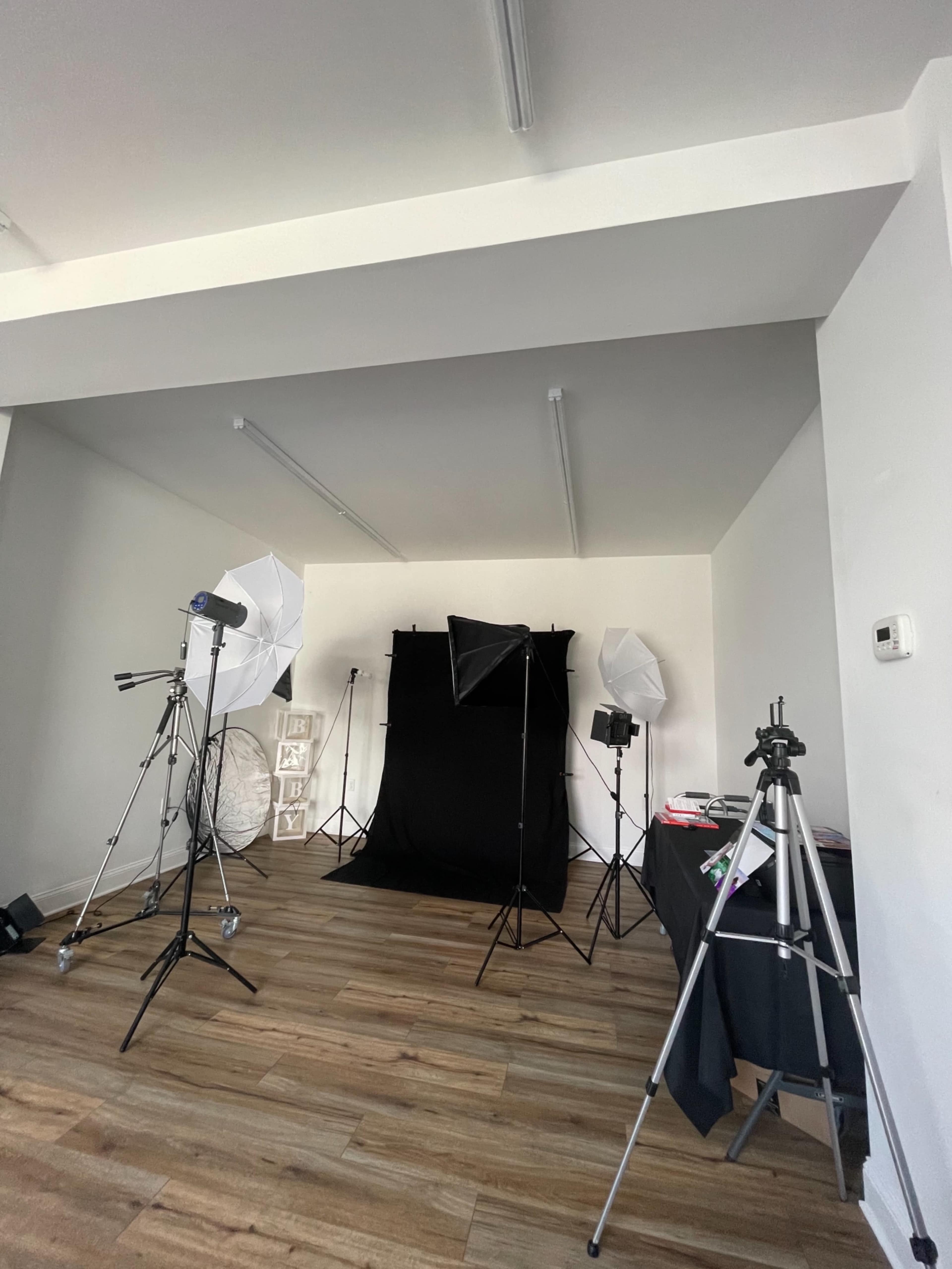 A photography studio setup with a black backdrop, lighting equipment, and tripods arranged in a spacious room with wooden flooring.