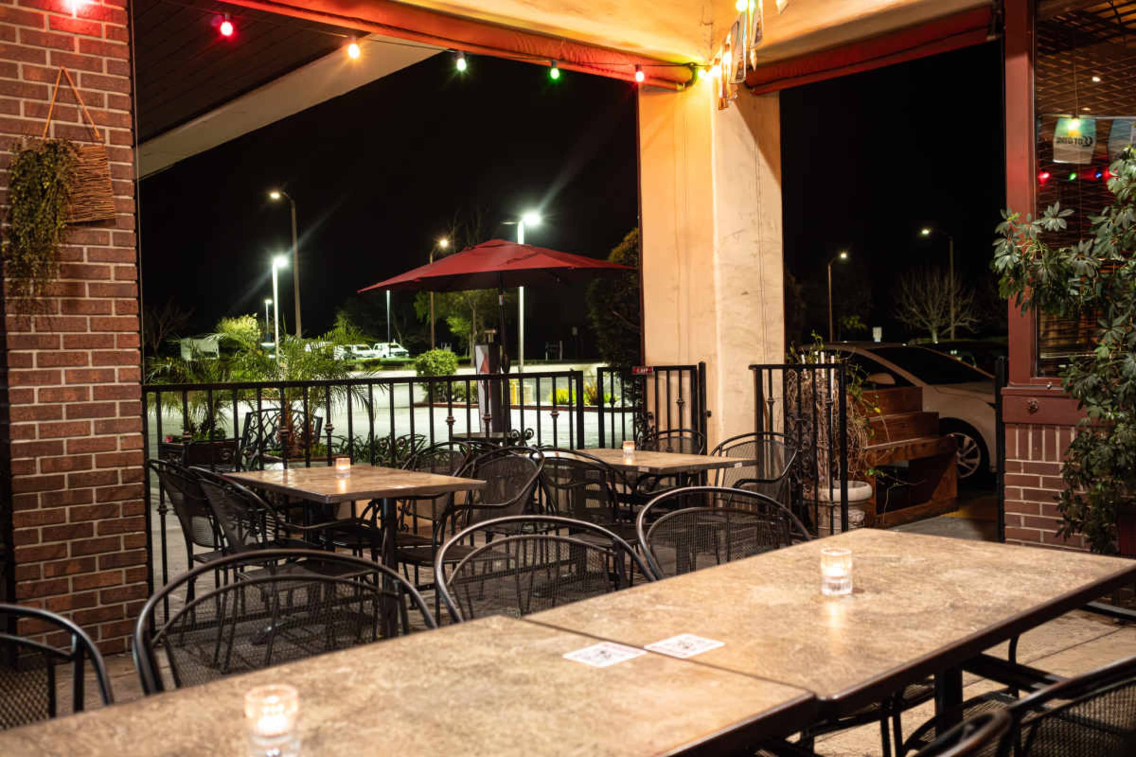 The image shows an empty outdoor dining area with several tables and chairs, illuminated by string lights at night.