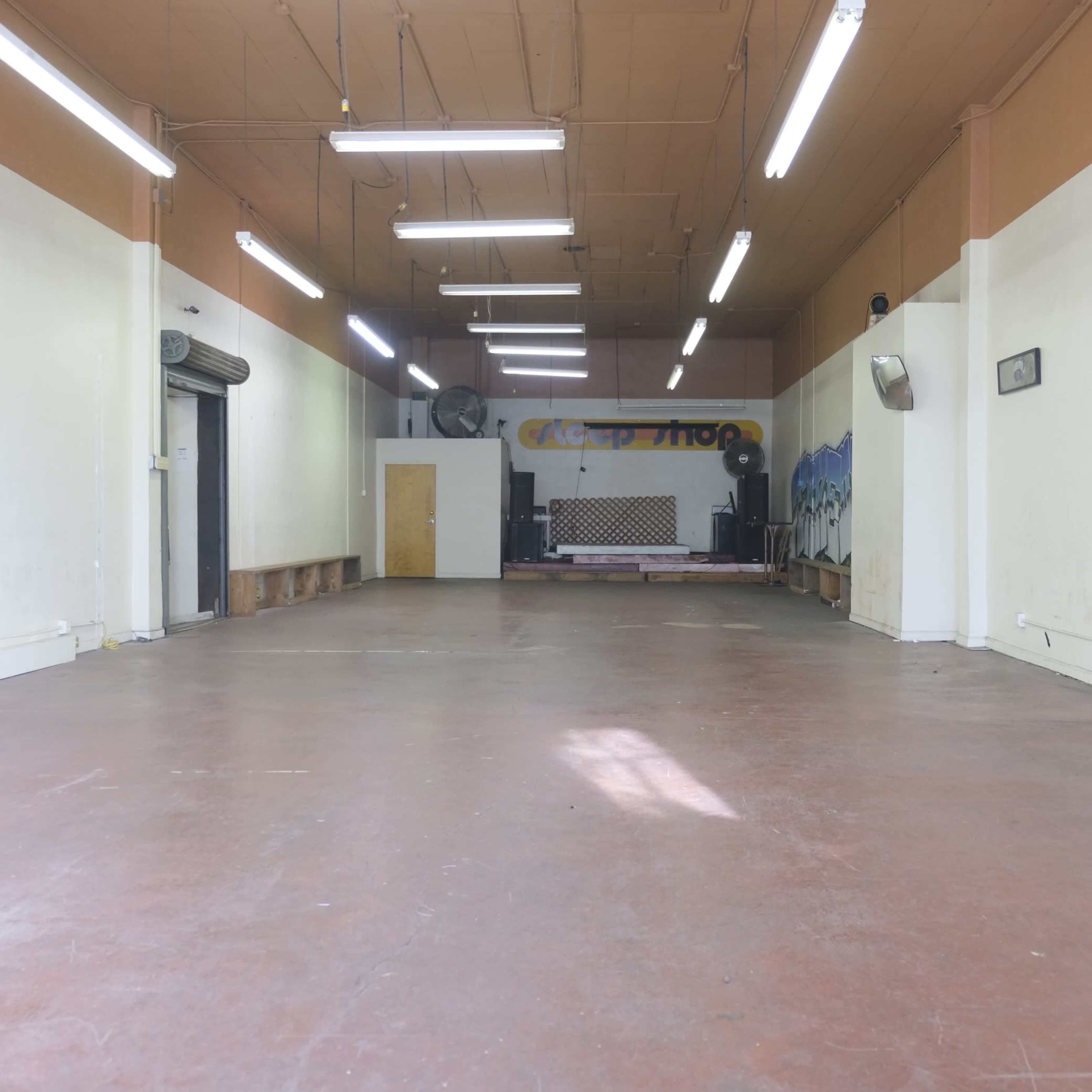 The image shows a spacious, empty room with a polished concrete floor, fluorescent lighting, and a stage area at the back.