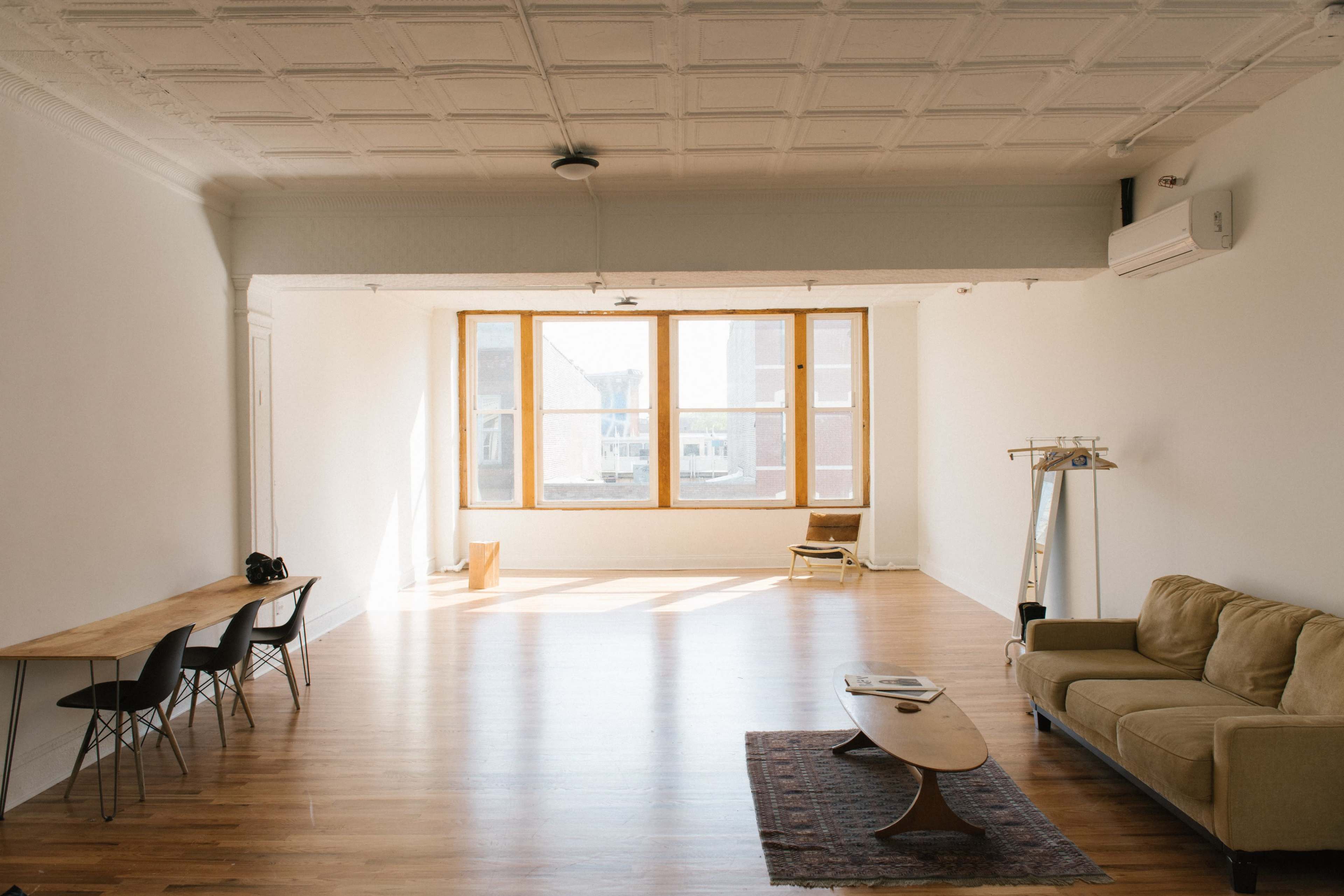 A spacious, brightly lit room with a wooden floor, large windows, a beige sofa, a low coffee table, and a dining area with black chairs.