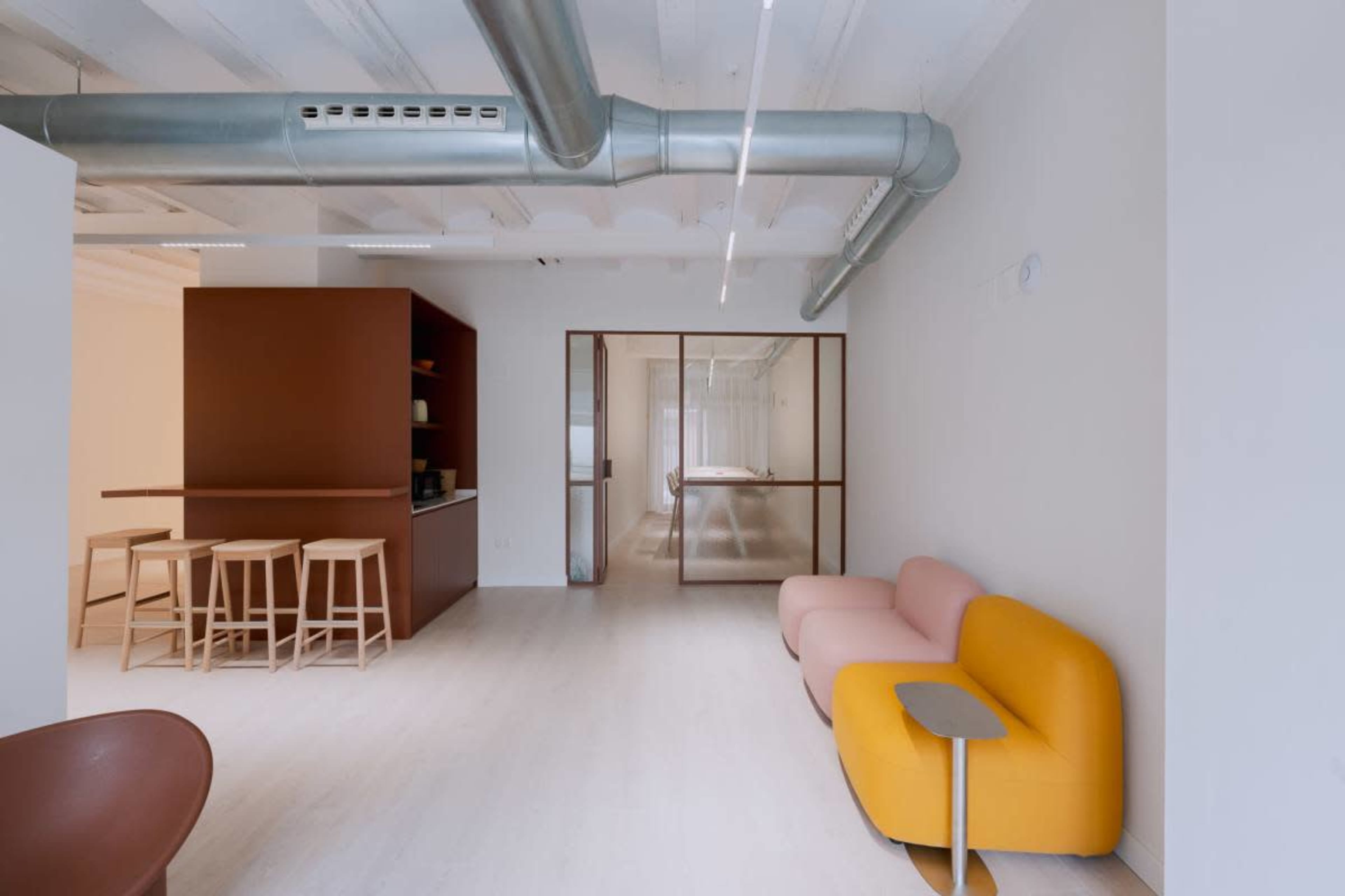 The image shows a modern office interior featuring a pink and yellow sofa, a wooden bar with stools, and a glass partition leading to a meeting room.