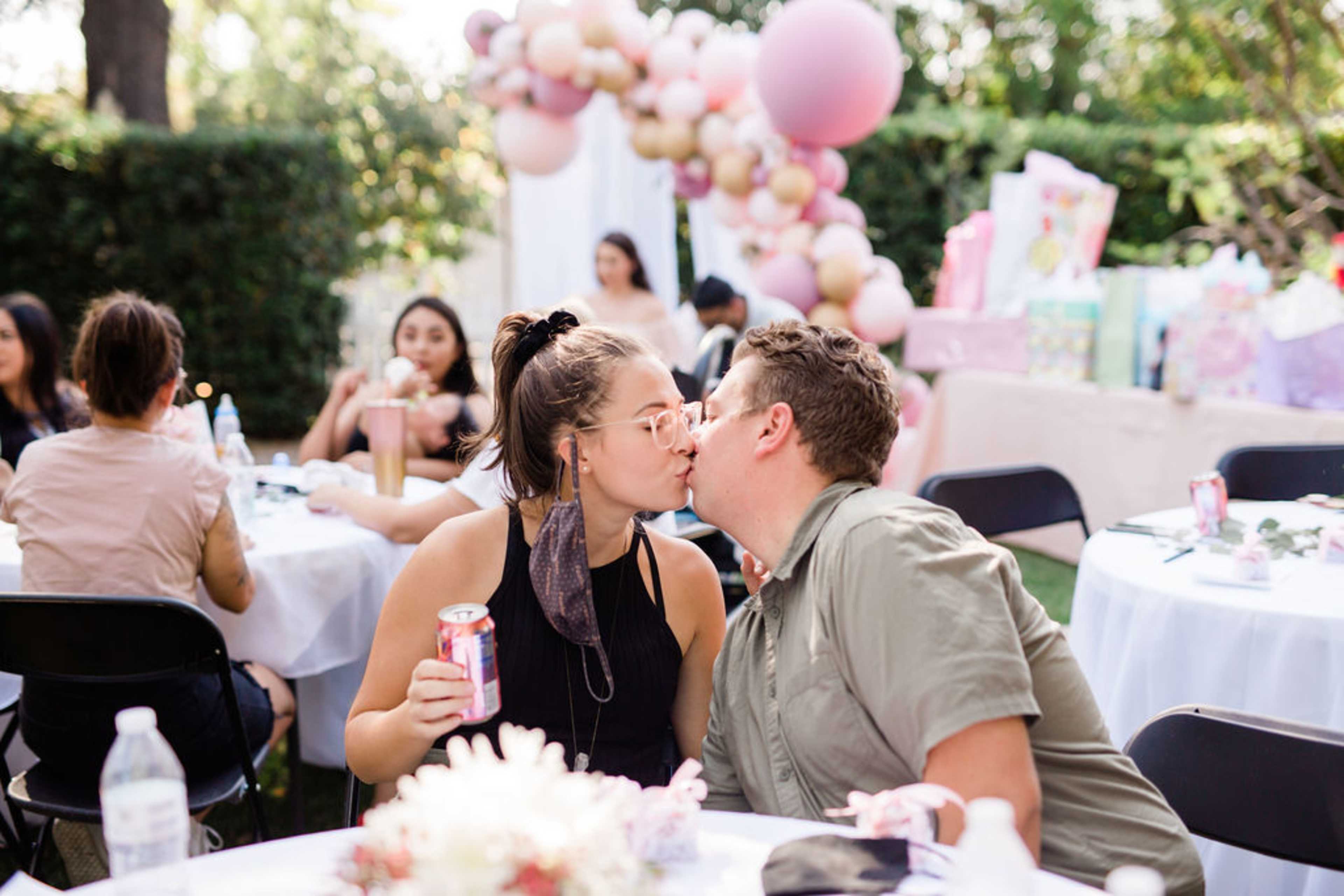 A couple shares a kiss at a celebration with balloons and gifts in the background while seated at a table.