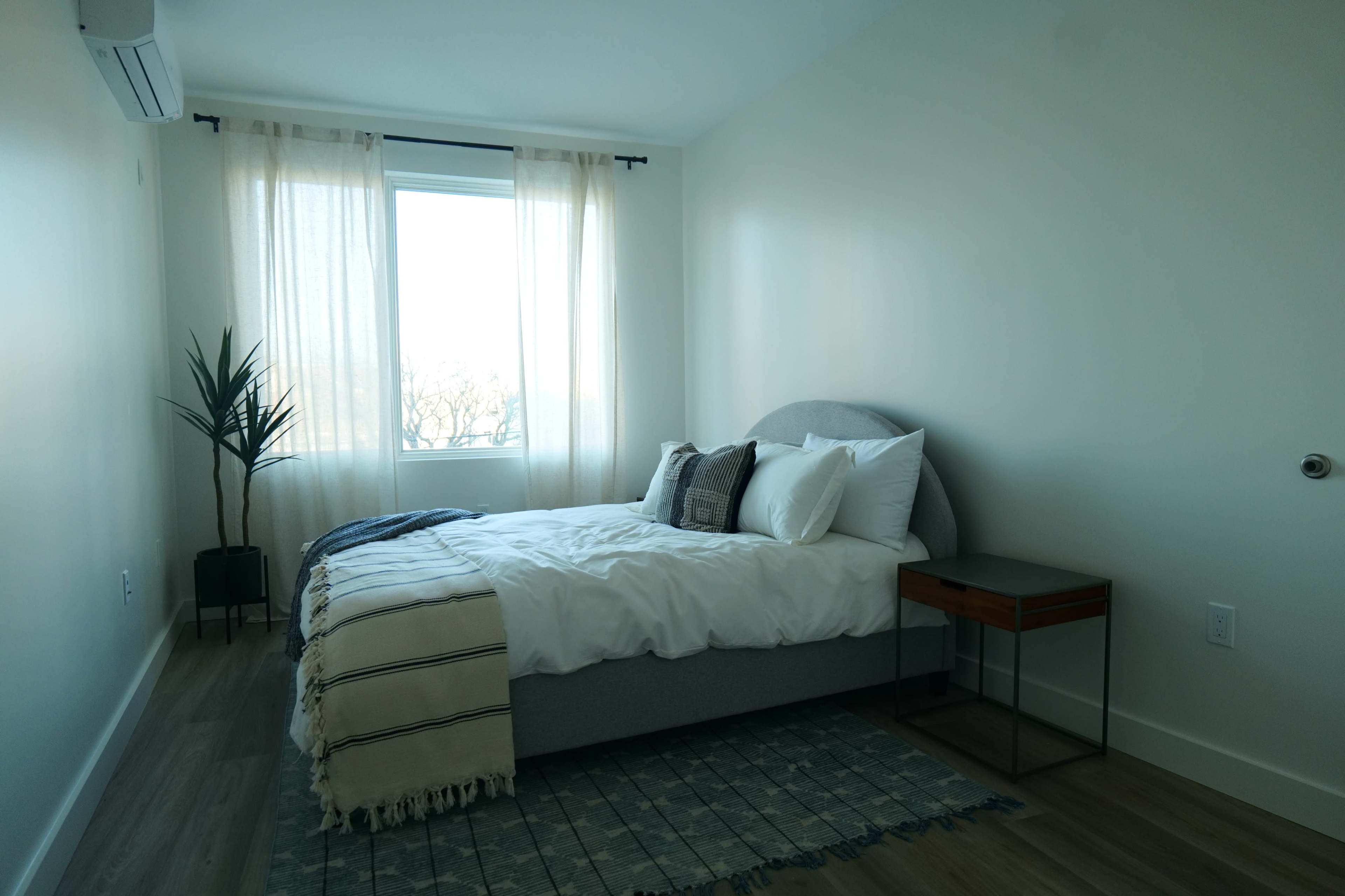 A minimalist bedroom featuring a bed with white linens, a small nightstand, and a window with sheer curtains.