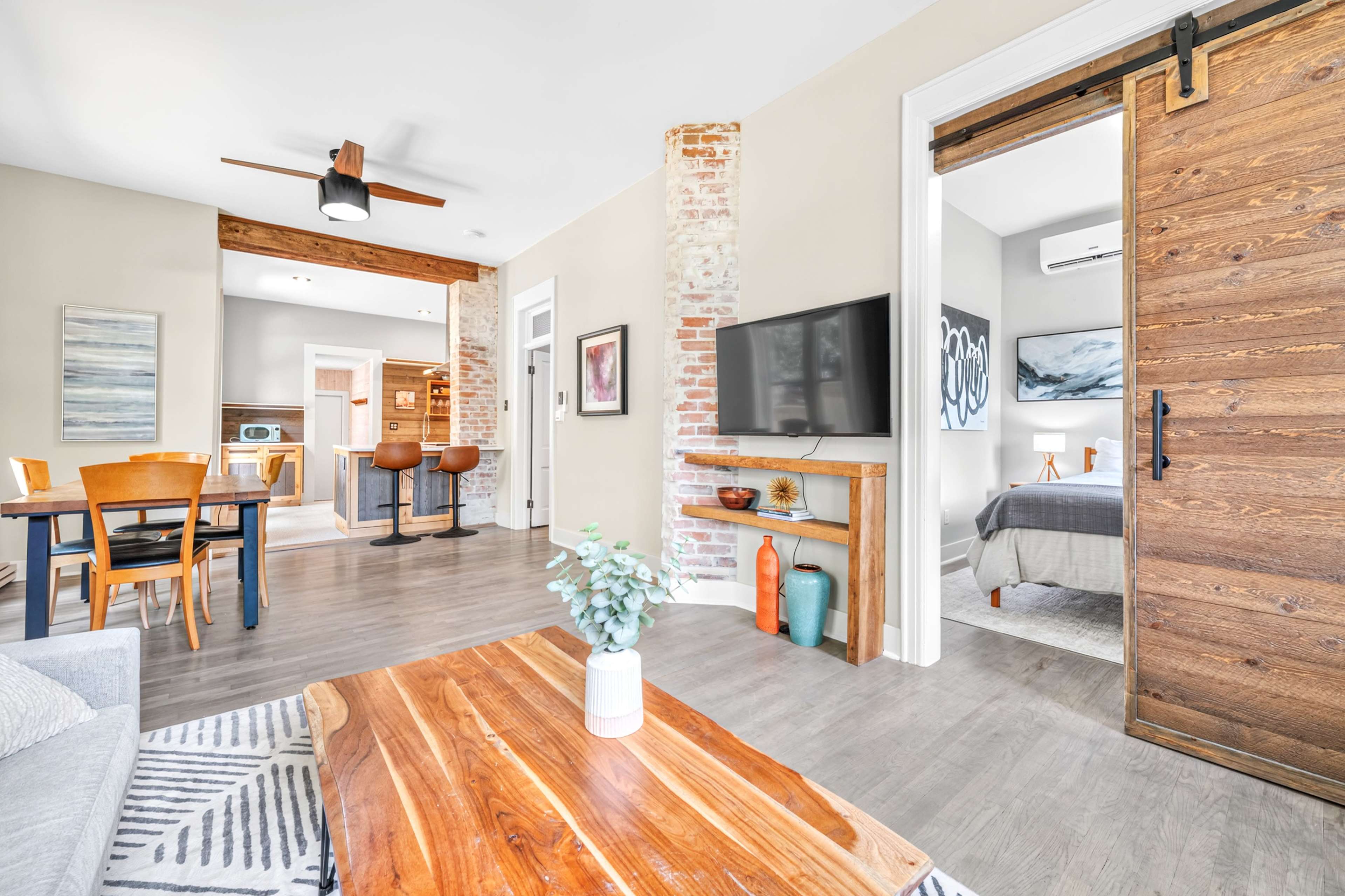 The image shows a modern open-concept living space featuring a seating area, dining table, and a view into a bedroom with a sliding barn door.