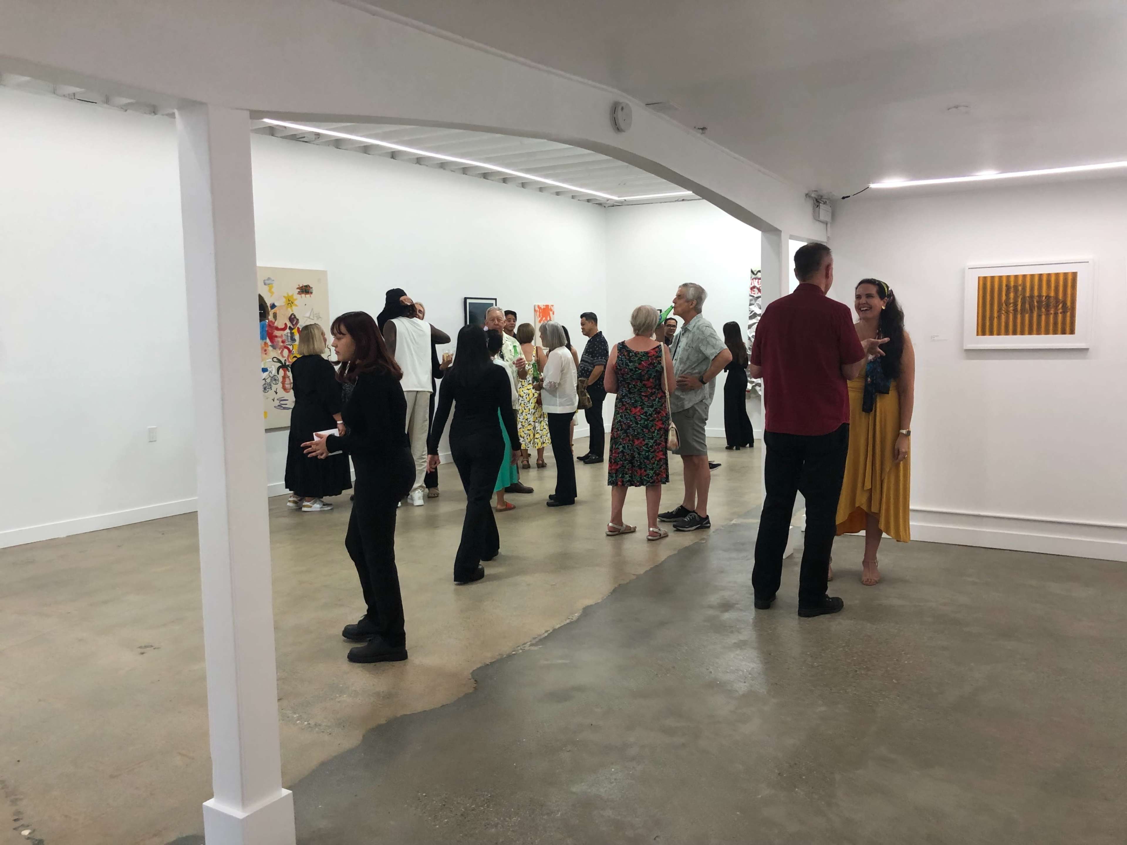 A group of people is gathered in a spacious art gallery, engaging in conversation while observing various artworks on the walls.