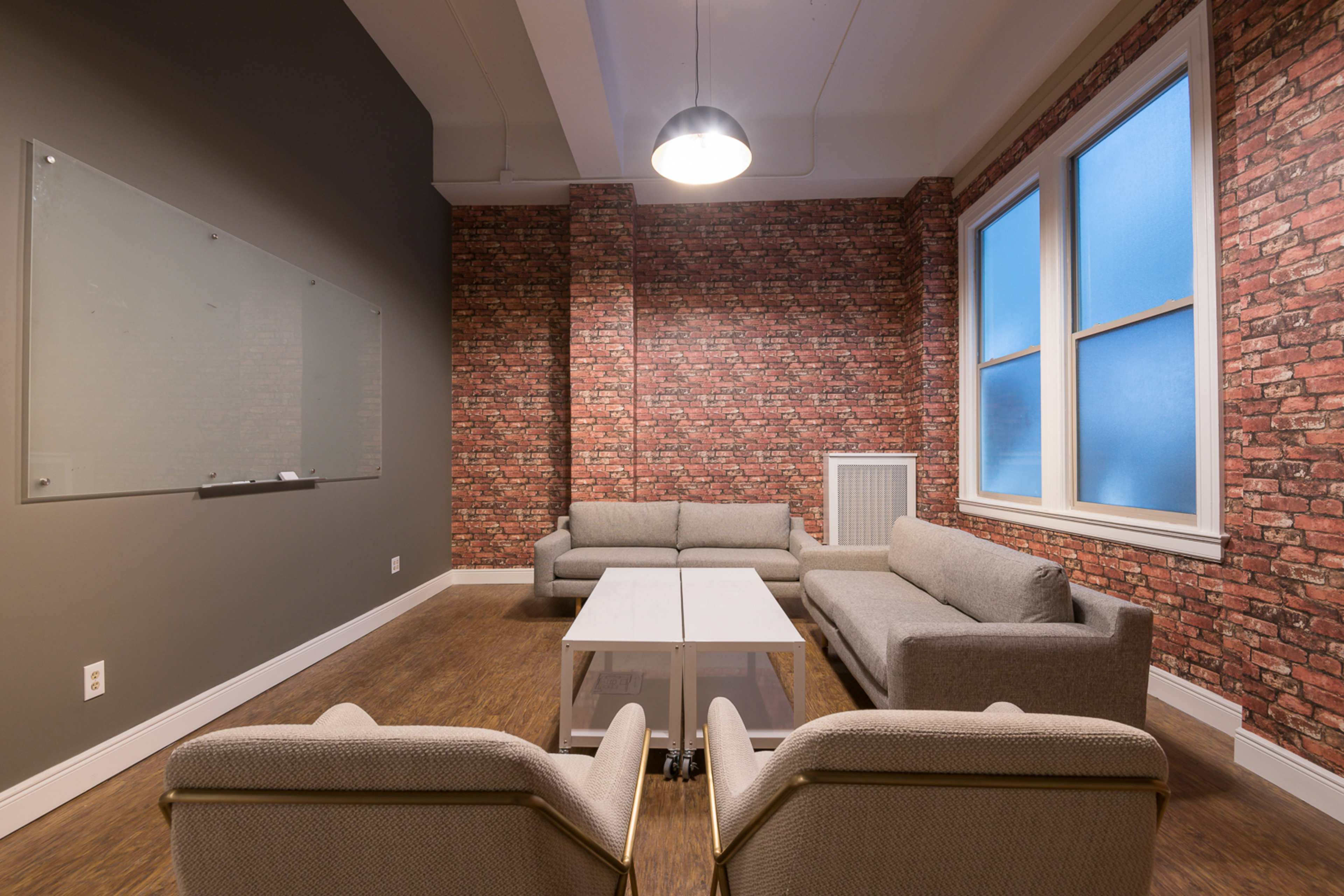 The image shows a modern lounge area with two gray sofas, two armchairs, and a glass coffee table, set against a wall featuring a brick pattern.