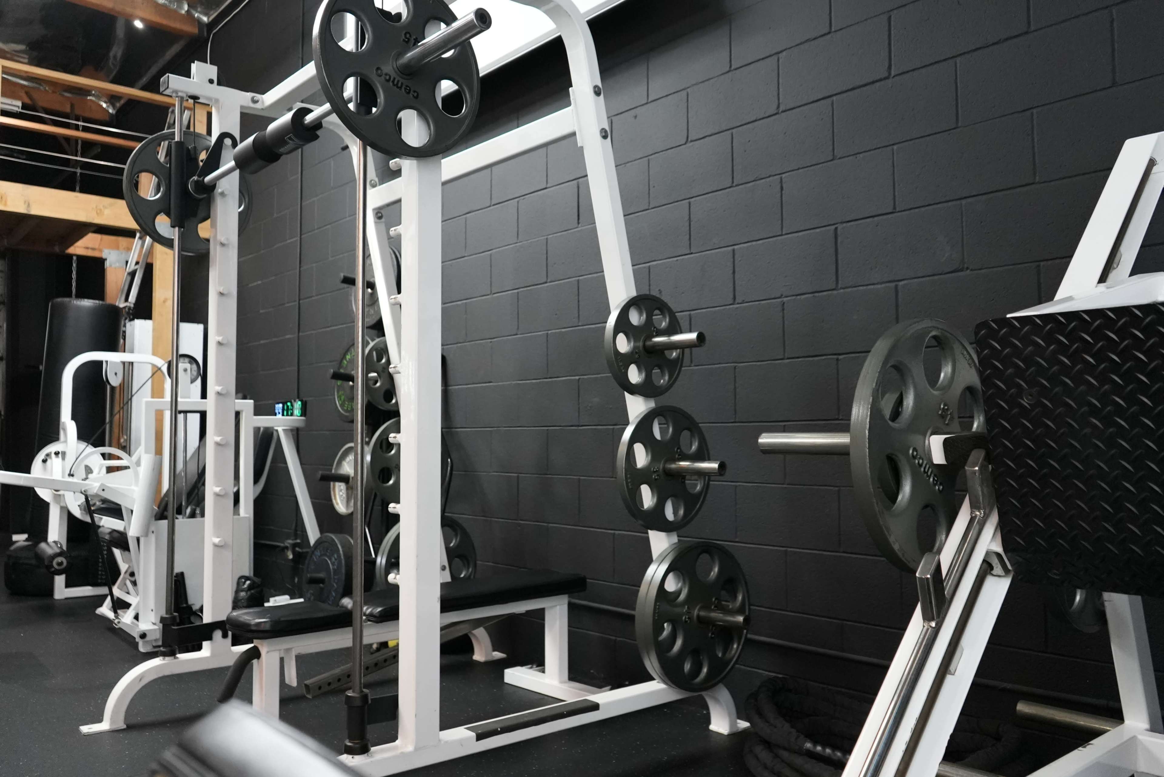 A fitness gym interior with various weightlifting equipment, including a barbell rack and weight plates on a black wall backdrop.