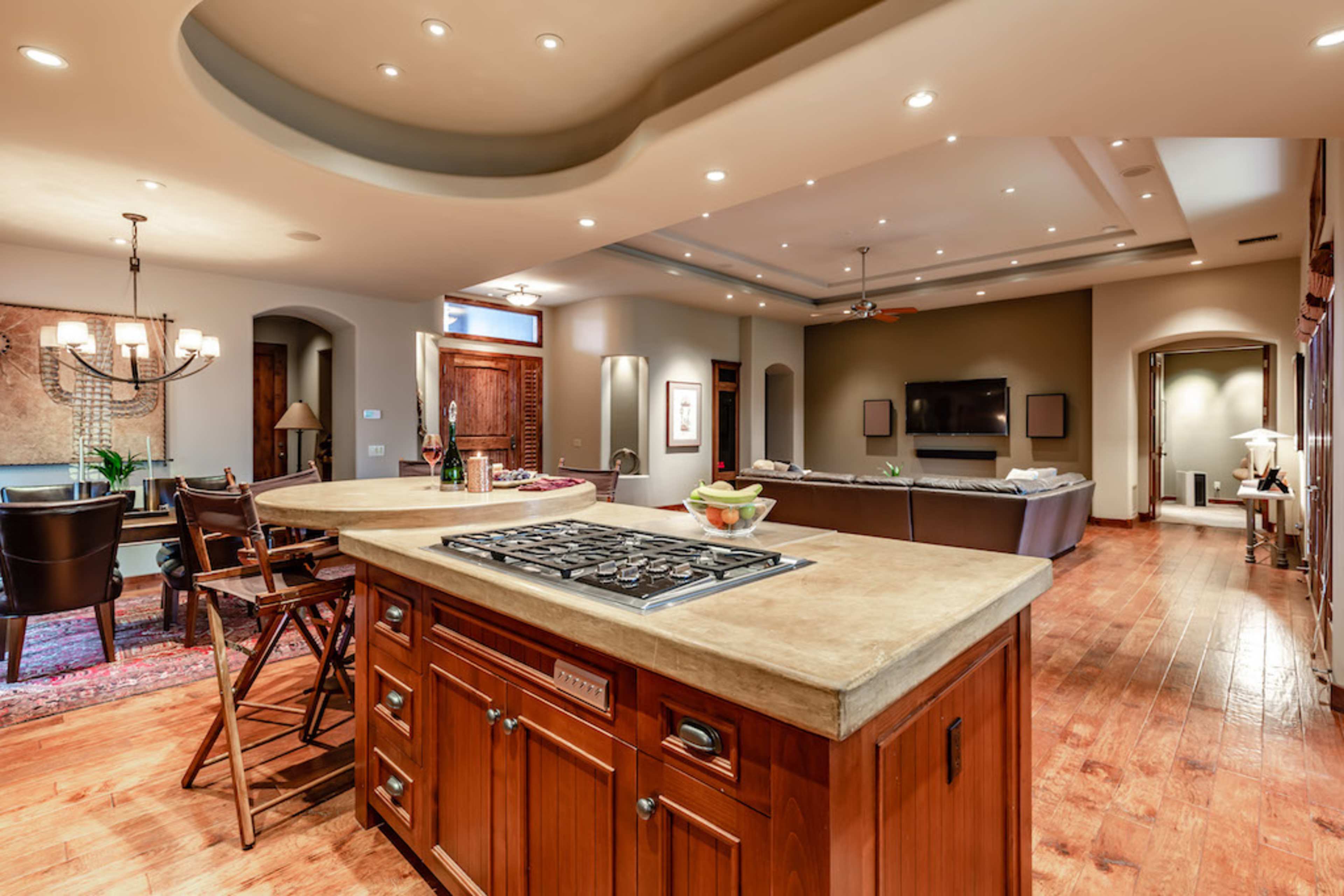 A spacious kitchen features a central island with a stovetop, surrounded by dining and living areas with wooden flooring and recessed lighting.