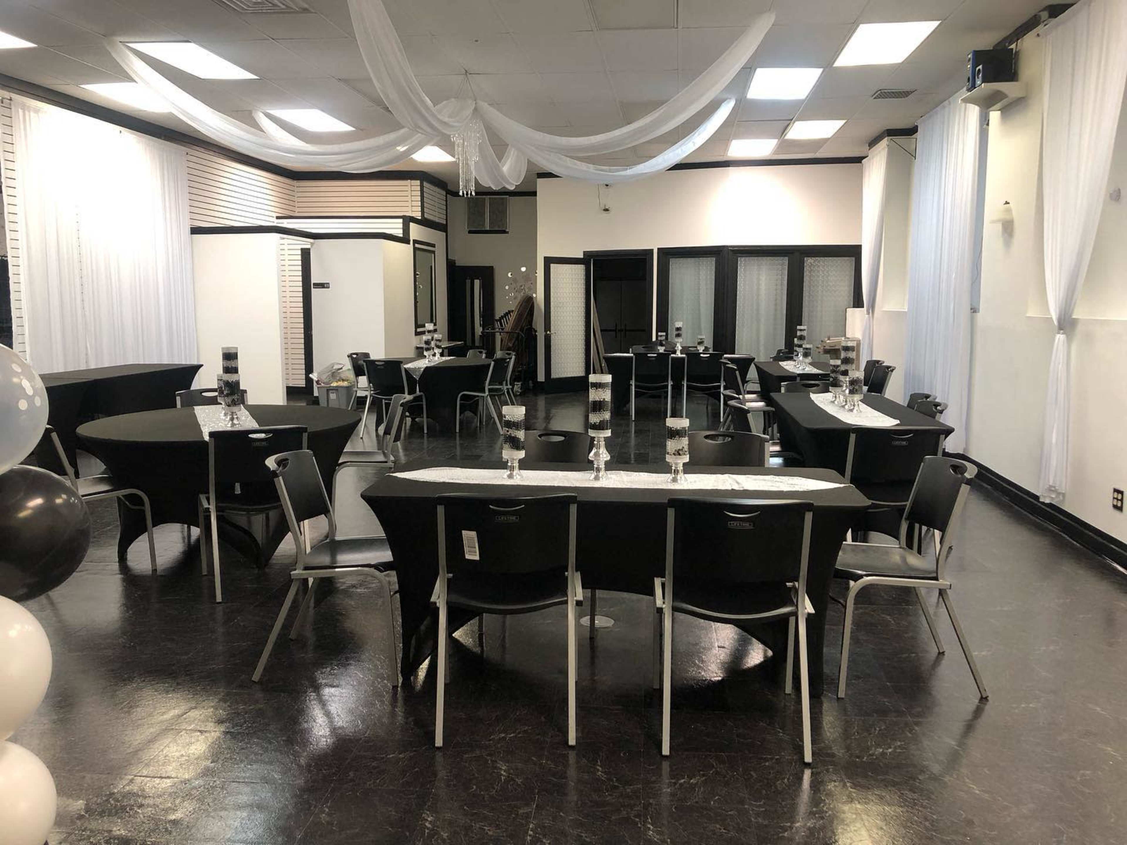 A large event space is set up with several round and rectangular tables, each adorned with tall glass centerpieces, and draped fabric hangs from the ceiling.