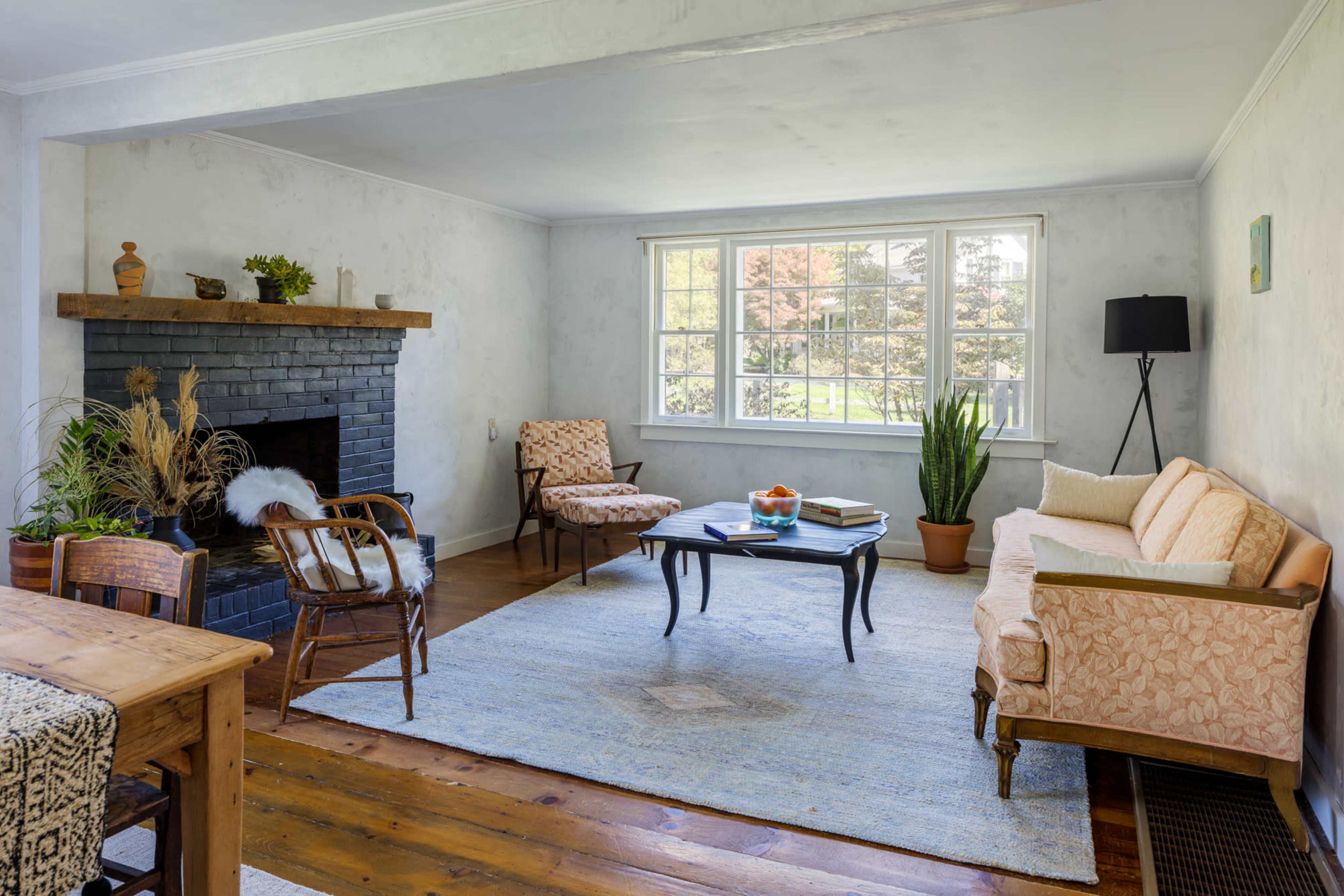 A living room features a sofa, two armchairs, a coffee table, and a fireplace, with large windows allowing natural light to enter.