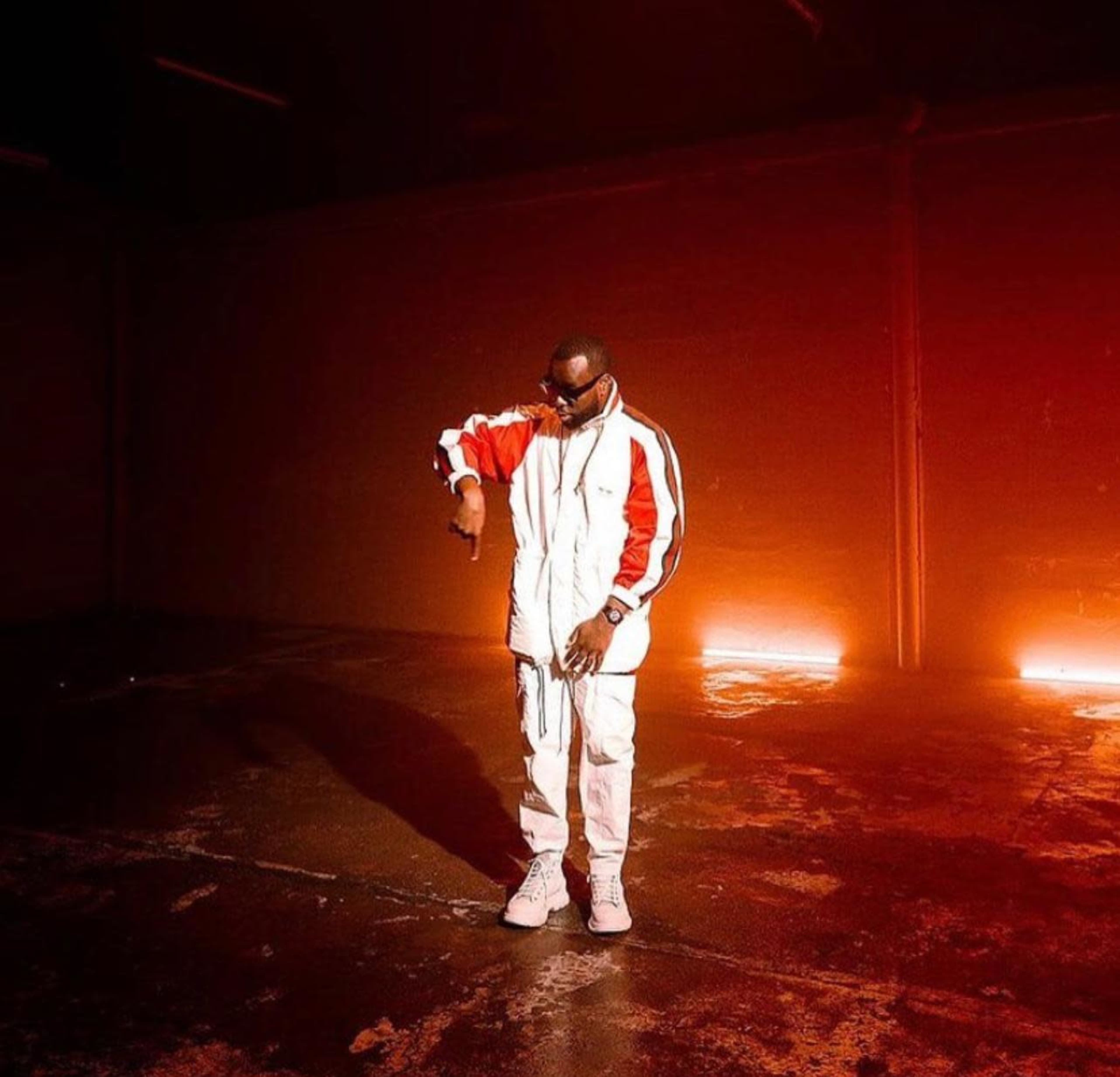 A man dressed in a white tracksuit with red sleeves stands in a dimly lit, empty warehouse space.