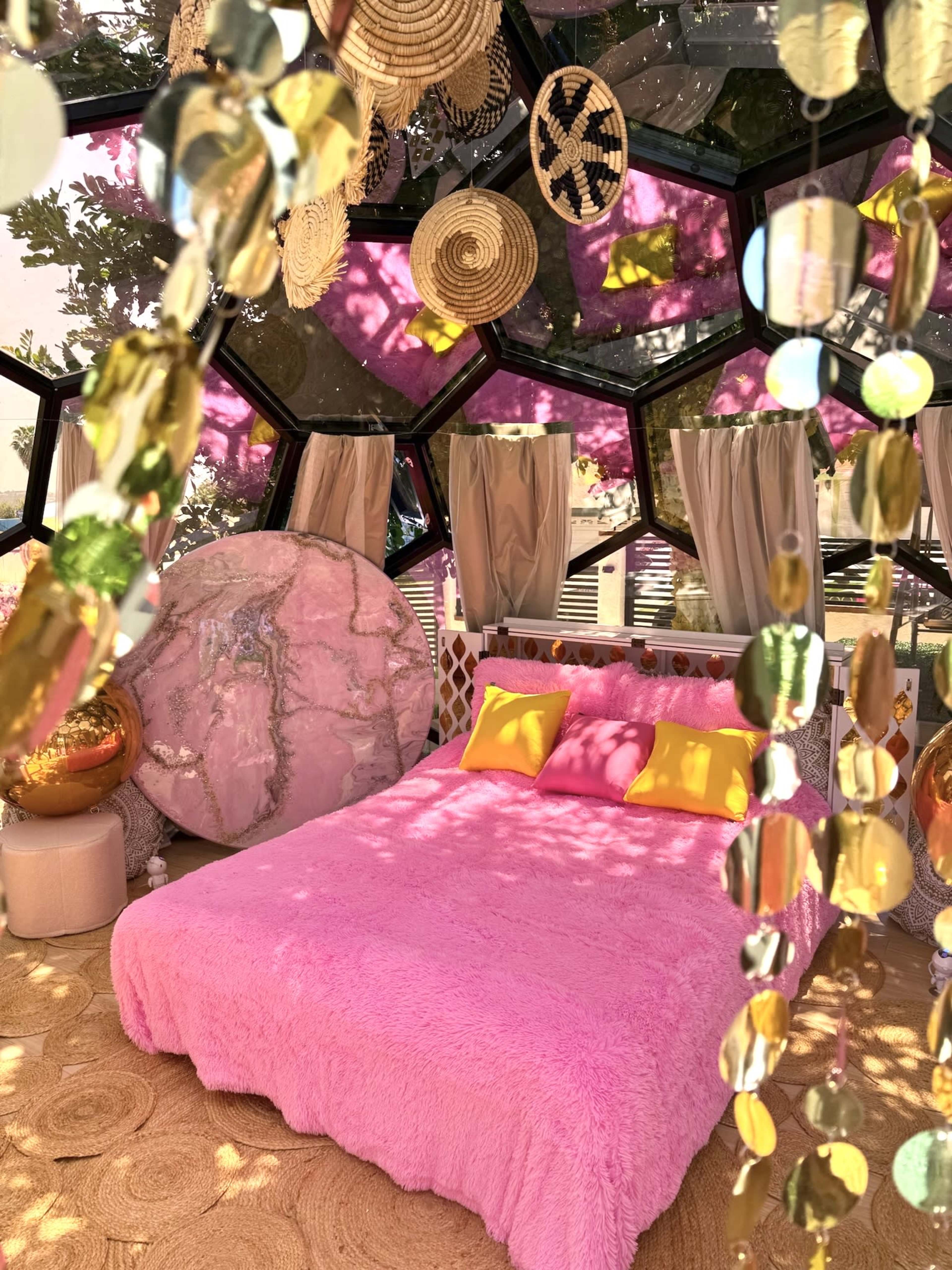 The image shows a brightly decorated geodesic dome interior with a pink bedspread, woven wall hangings, and decorative elements like bead curtains and cushions.