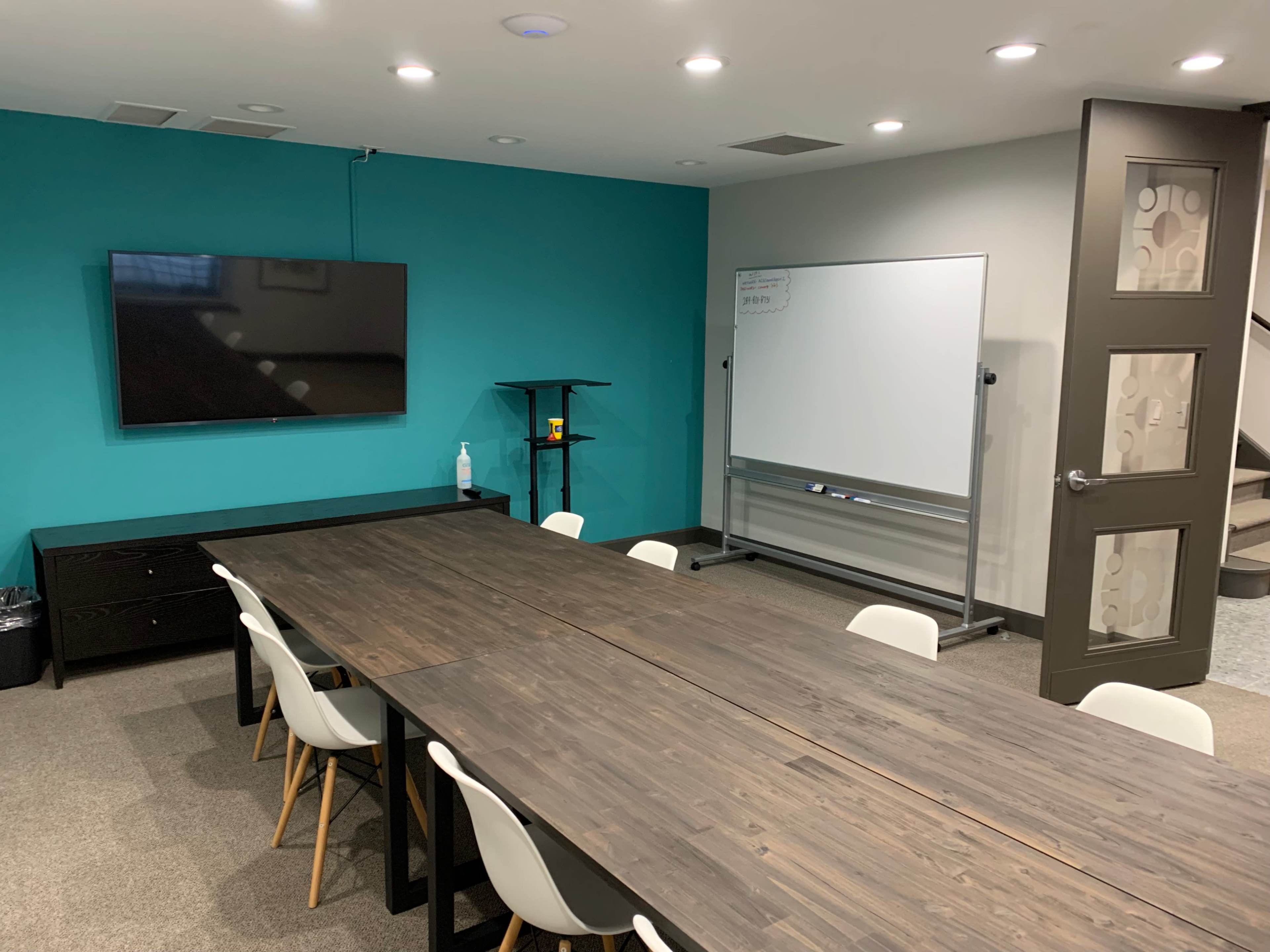 The image shows a conference room with a large wooden table, white chairs, a wall-mounted TV, and a whiteboard on a blue wall.