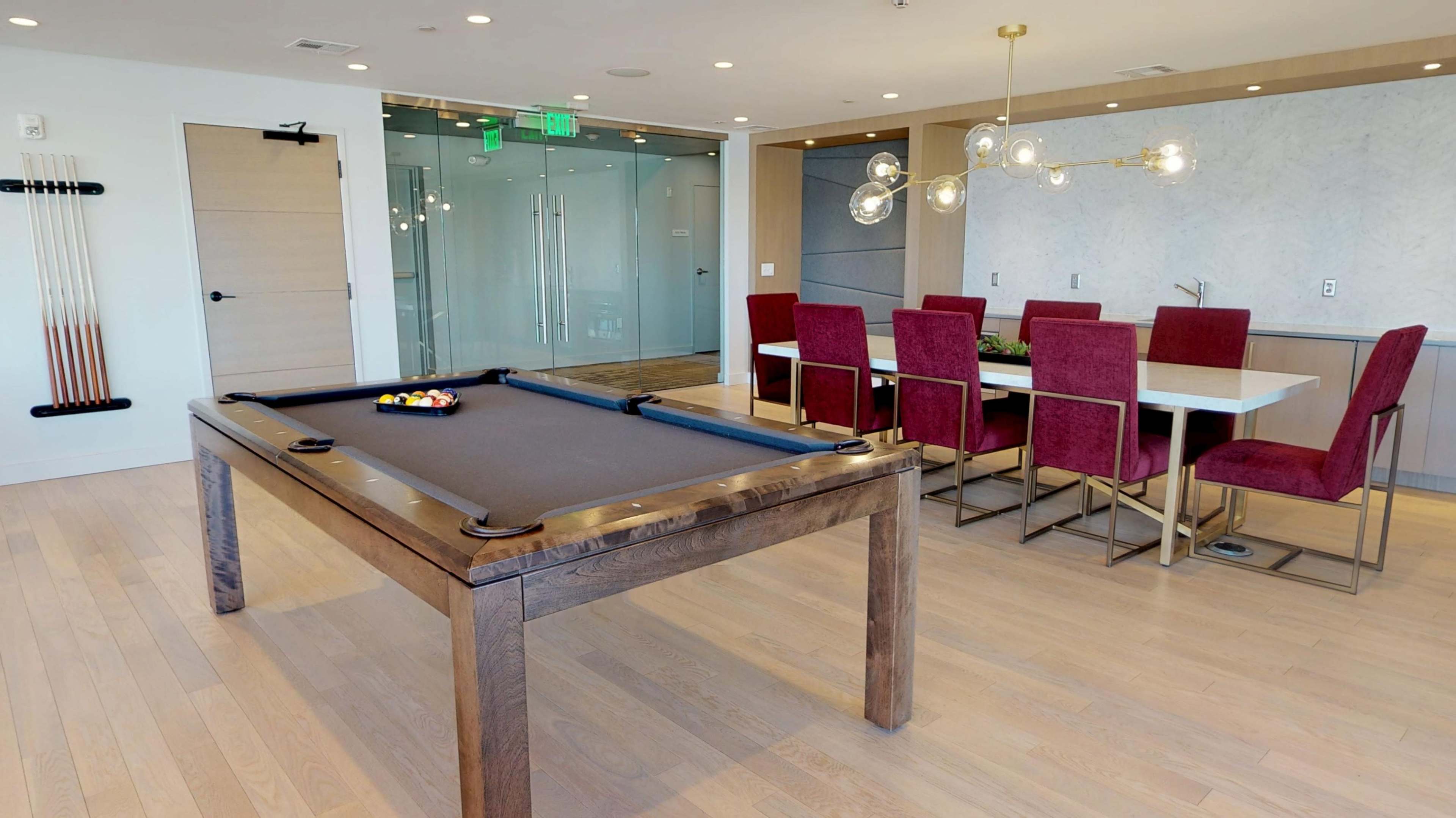 The image shows a modern lounge area featuring a billiard table and a dining space with a long table surrounded by red chairs.