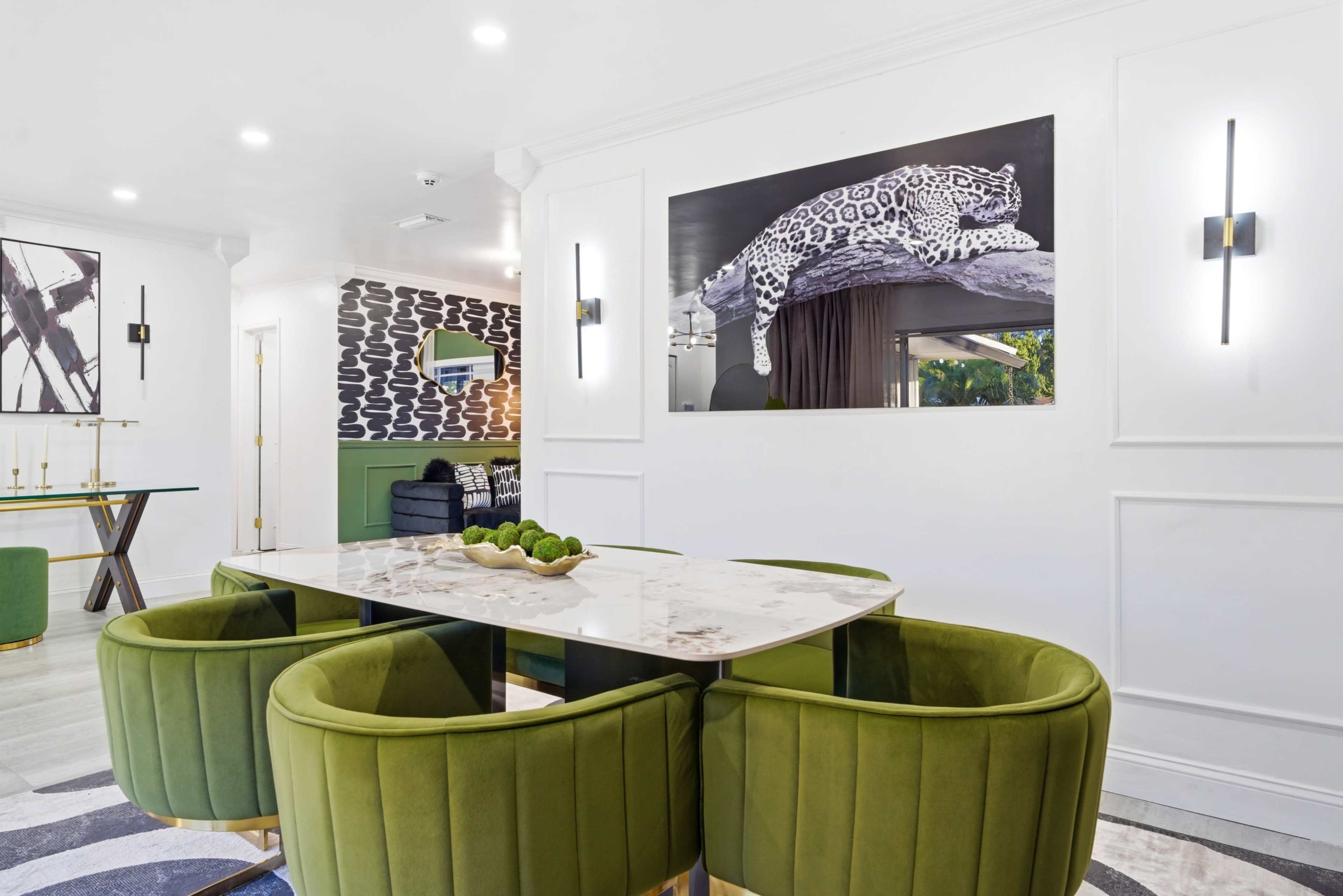 The image displays a modern dining area featuring a marble table surrounded by green velvet chairs, with a large print of a leopard on the wall behind.