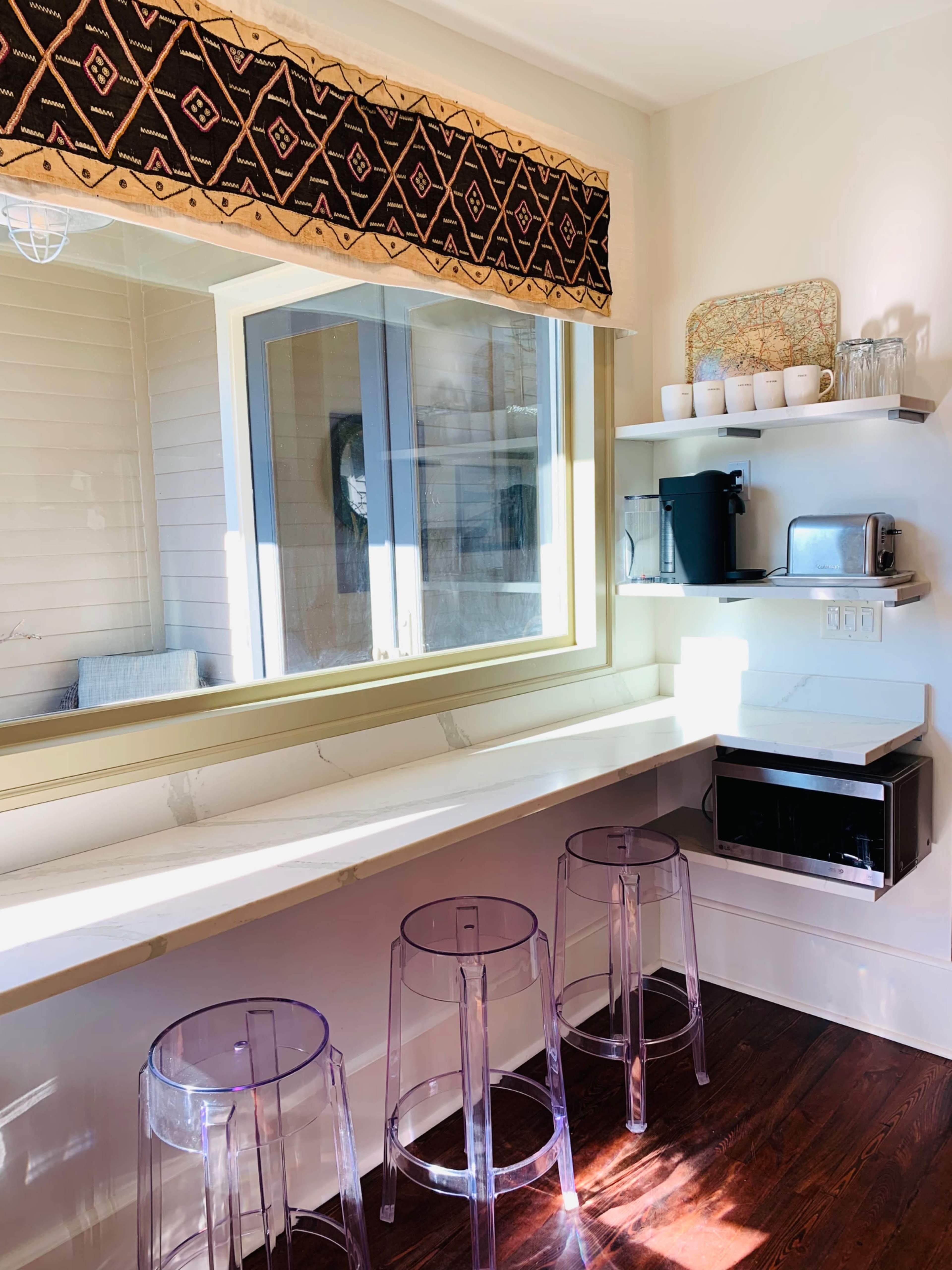 A modern kitchen area with a long countertop, three transparent stools, and shelves displaying various kitchen items.