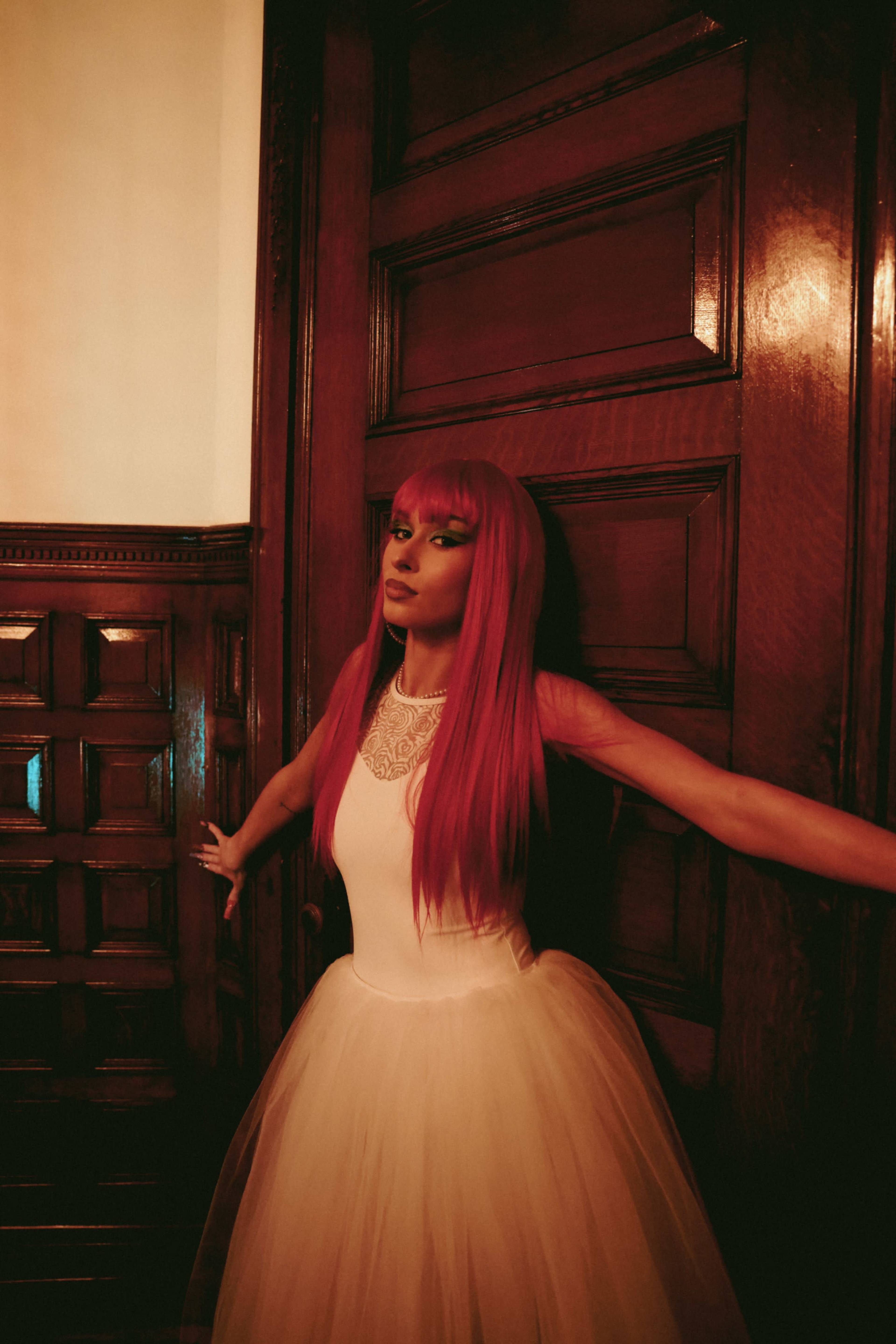 A person with long pink hair stands against a wooden door, wearing a white dress with a tulle skirt.