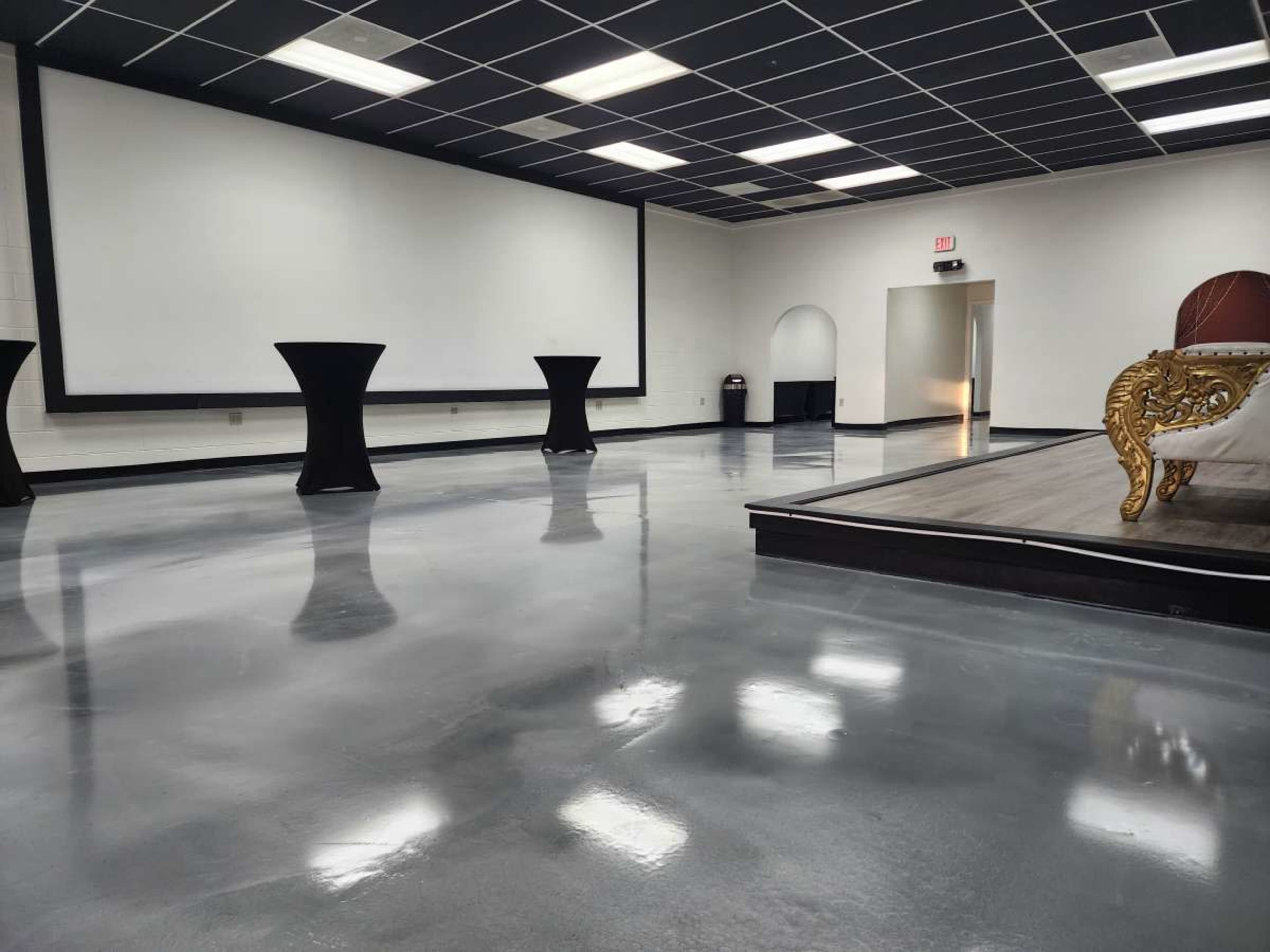The image shows a spacious, modern room featuring polished concrete floors, two tall round tables, a large projection screen, and an ornate seating area in the corner.