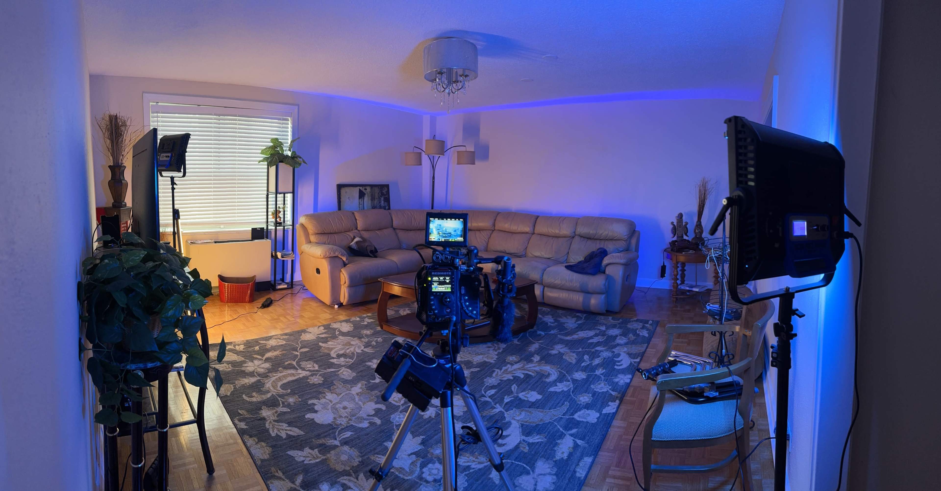 The image shows a spacious living room with a light-colored couch, a coffee table, two tripods with cameras, and various plants, all illuminated by blue lighting.