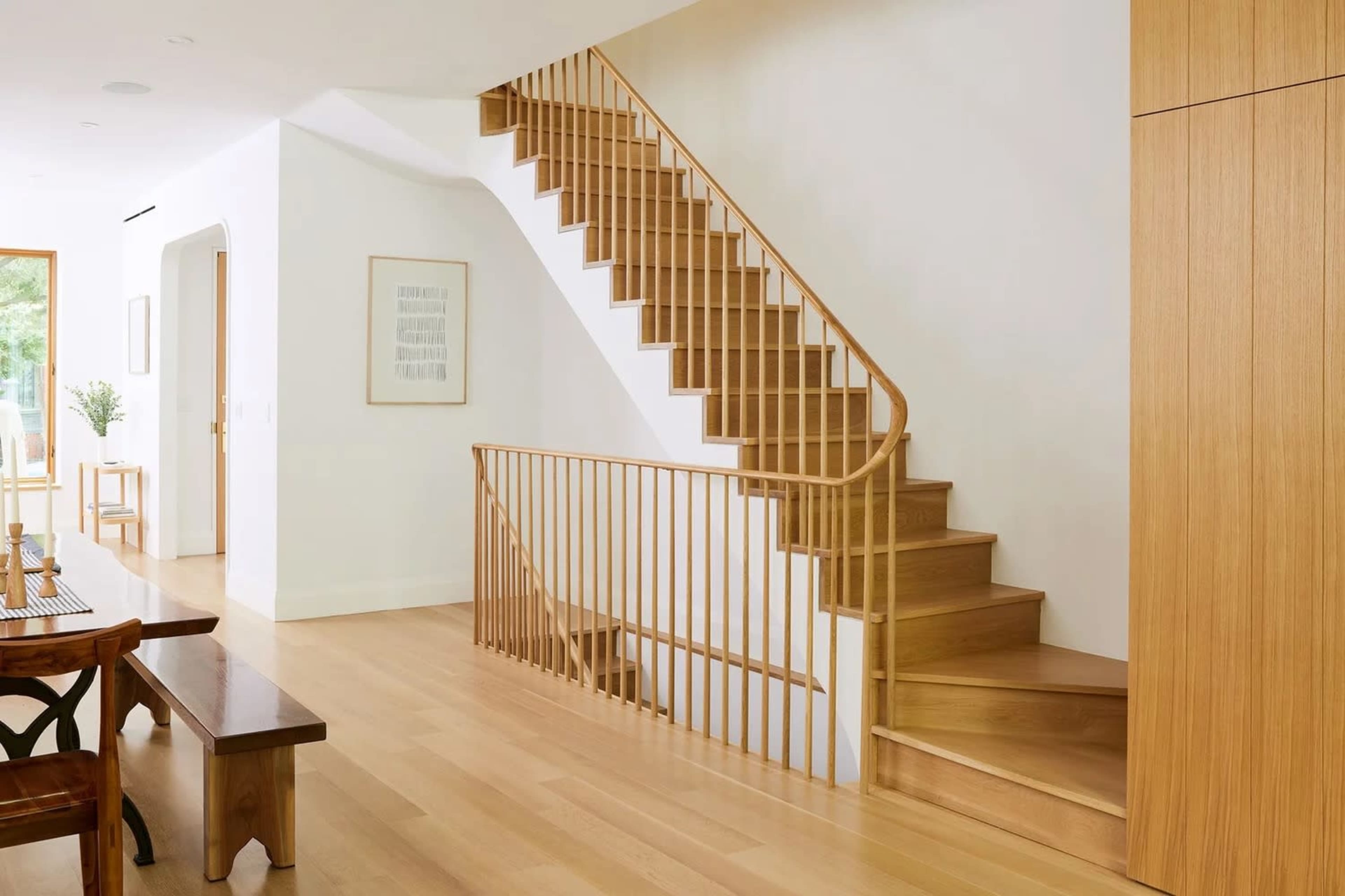 The image shows a modern interior space featuring a wooden staircase with a minimalist railing, hardwood floors, and light-colored walls.