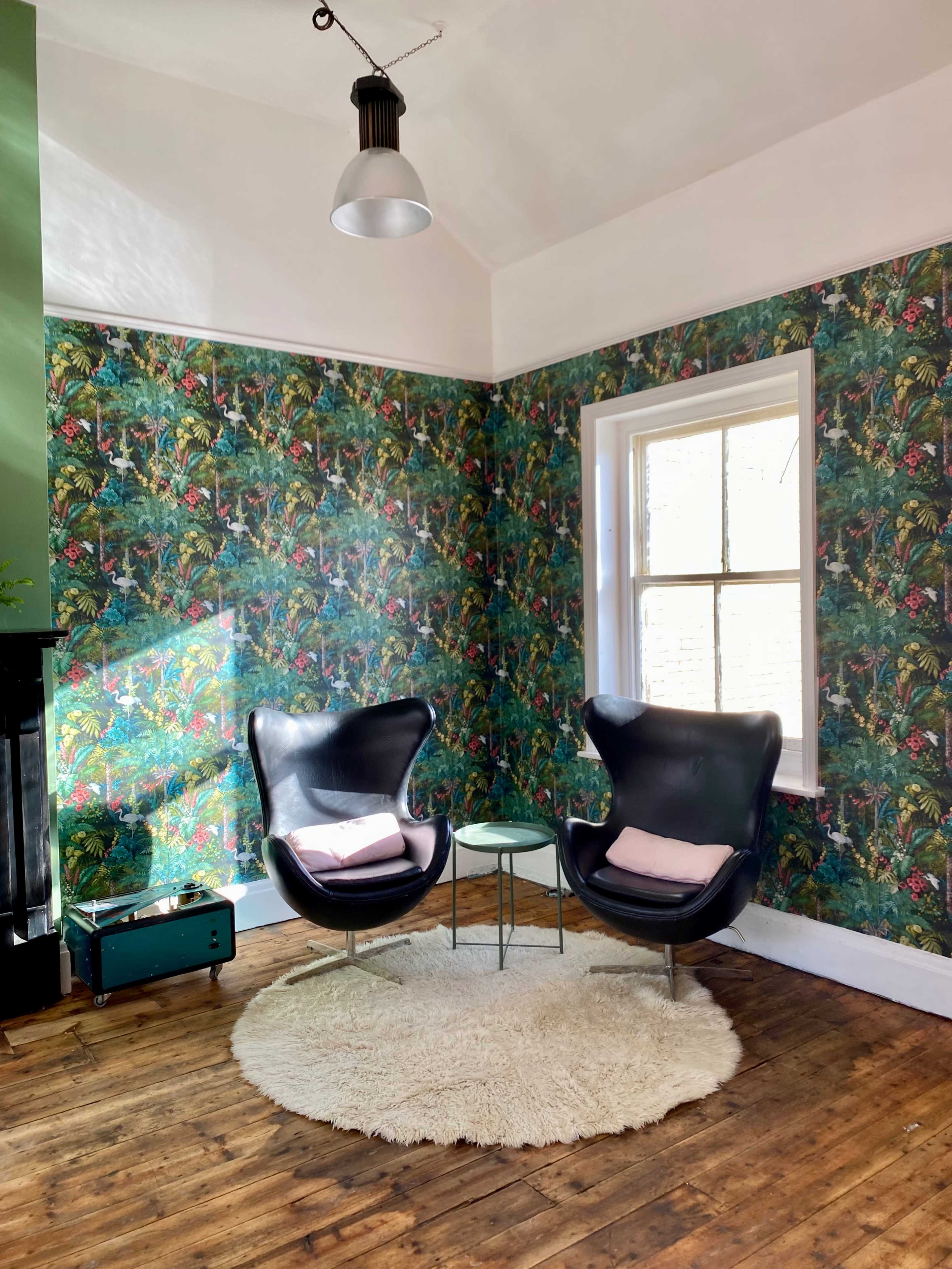 A cozy interior with two black chairs, a small glass table, and a light fixture, all set against a vibrant floral wallpaper.