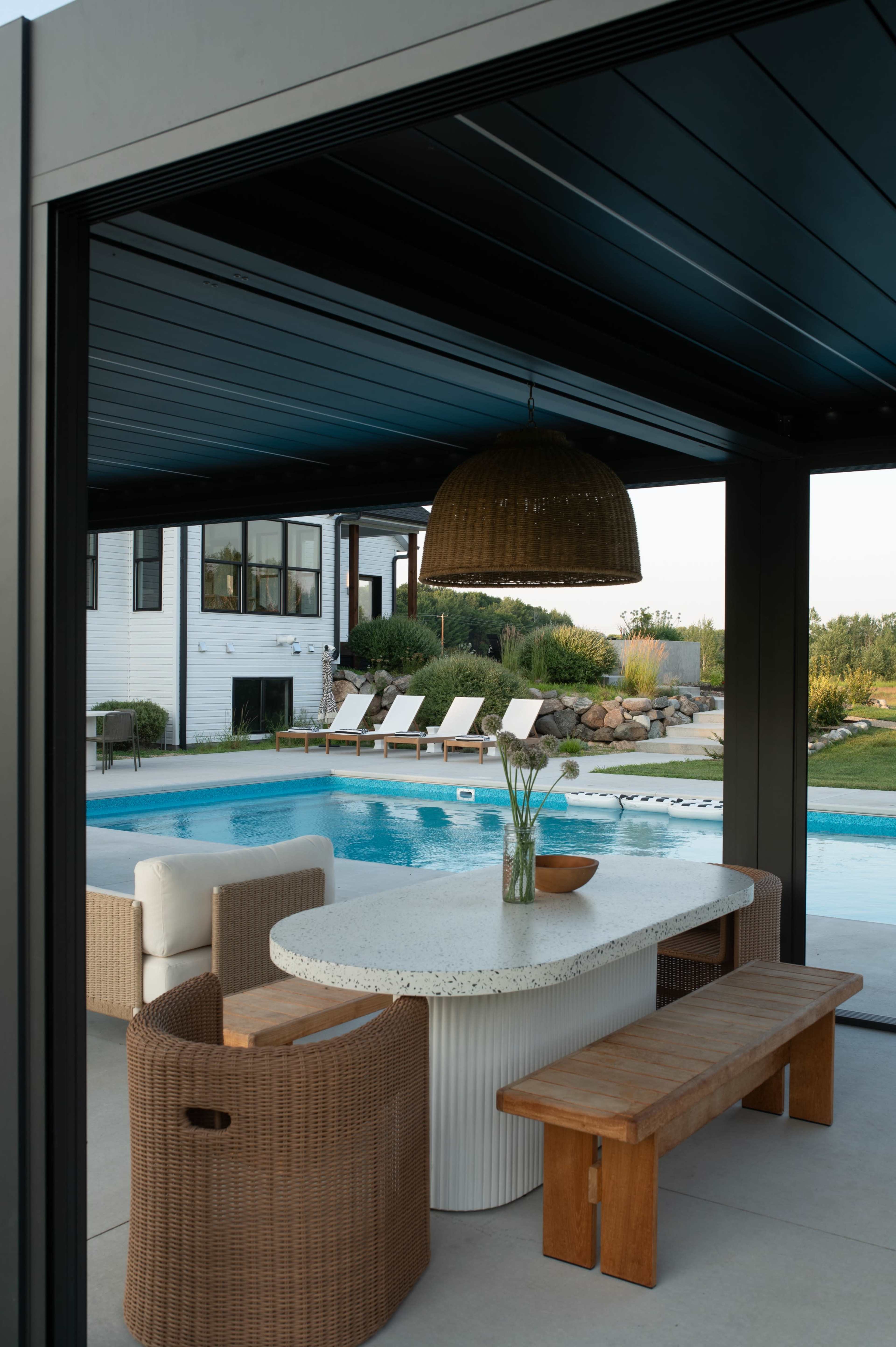 The image shows a modern outdoor dining area adjacent to a pool, featuring a round table with chairs and a wooden bench under a large pendant light.