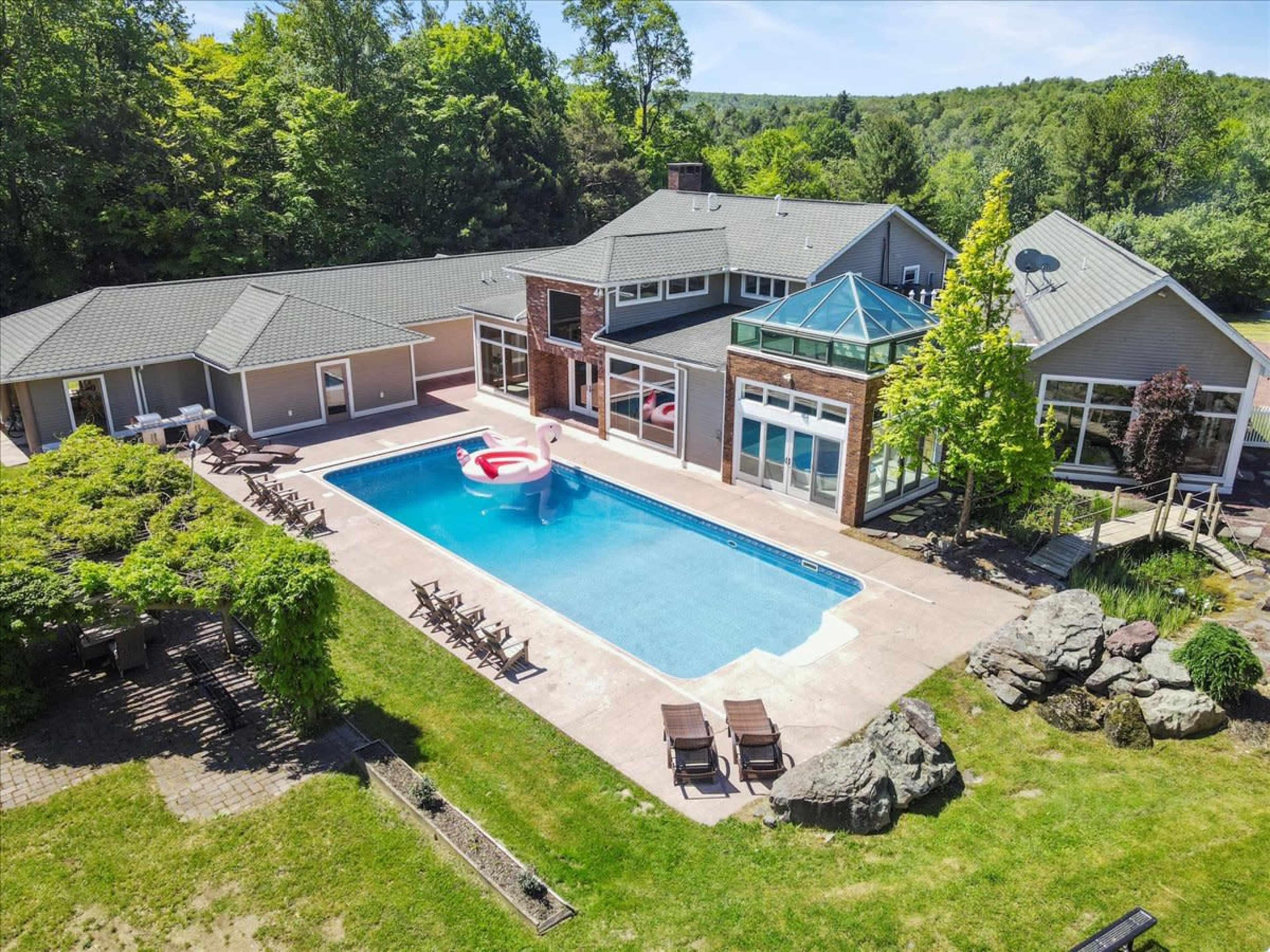 A modern house with large windows surrounds a rectangular swimming pool, featuring a giant inflatable flamingo, set against a backdrop of greenery.