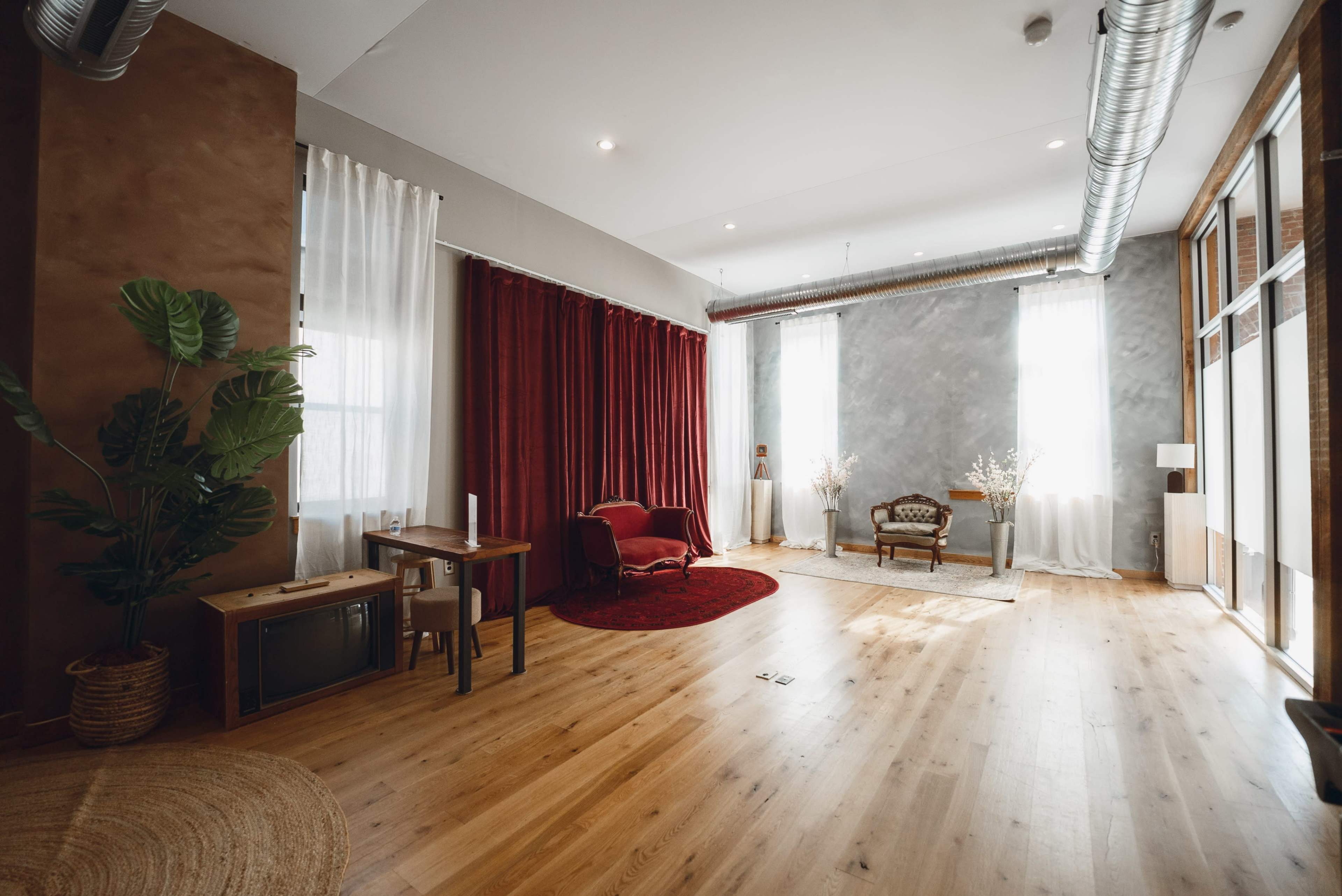 The image shows a spacious room with wooden flooring, large windows, and a mix of modern and vintage furnishings, including a red curtain, a small table, and a plush armchair.