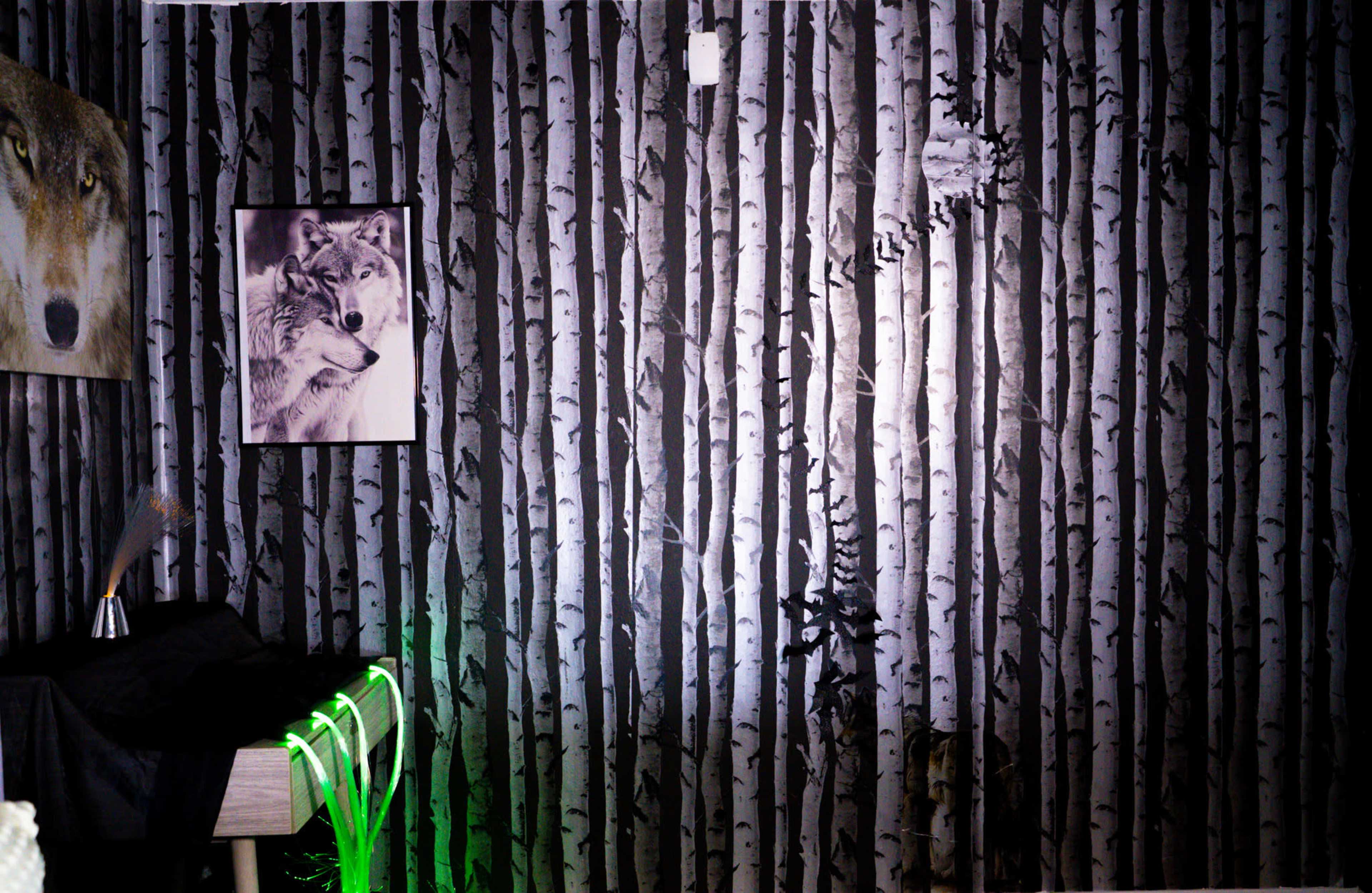 The image shows a room with wallpaper depicting black and white birch trees, a wolf portrait on the wall, and a table with illuminated green tubing.