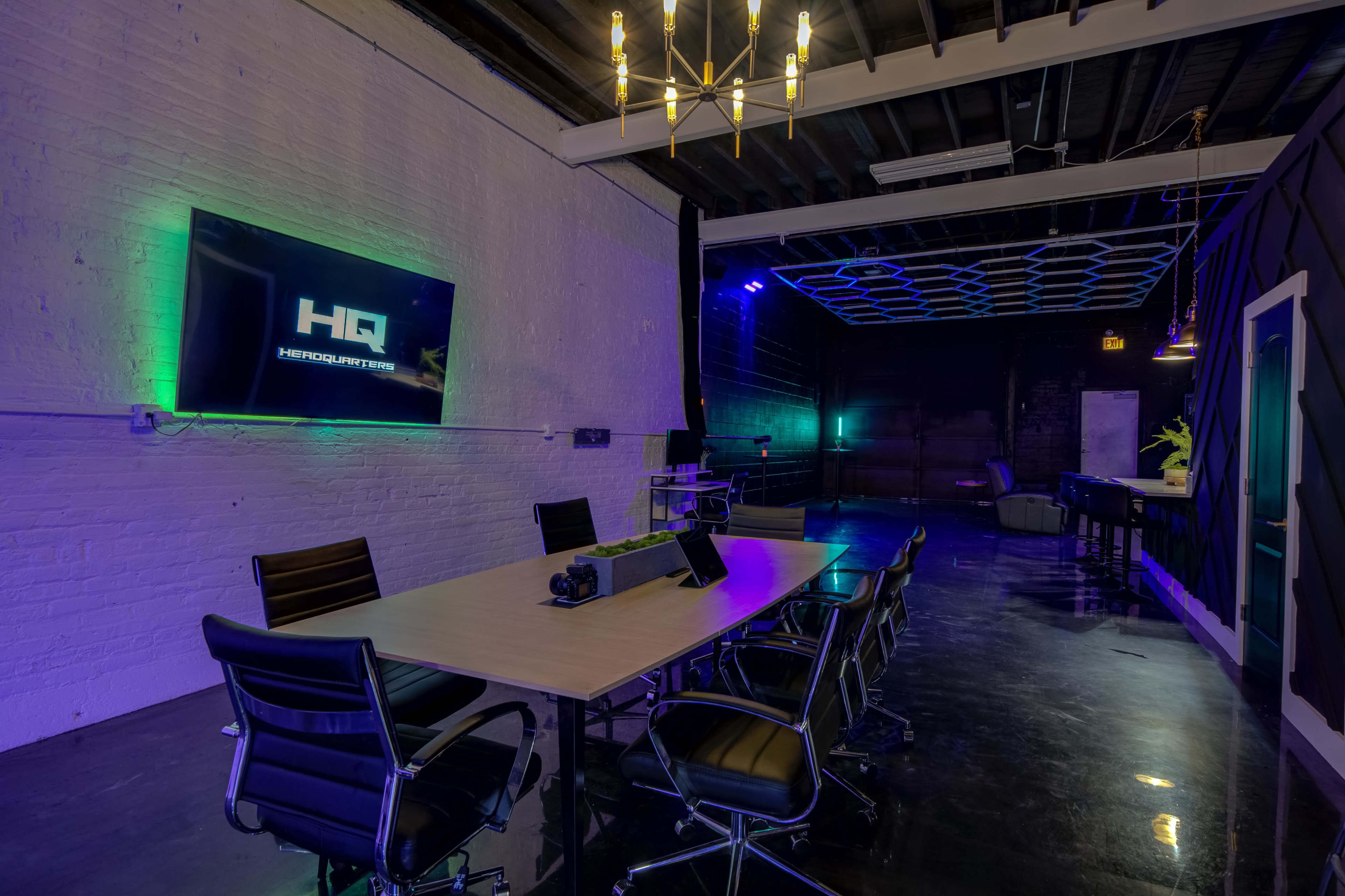 A modern conference room with a long table, black chairs, and a large screen displaying the letters "HQ" on a white brick wall, illuminated by various lights.