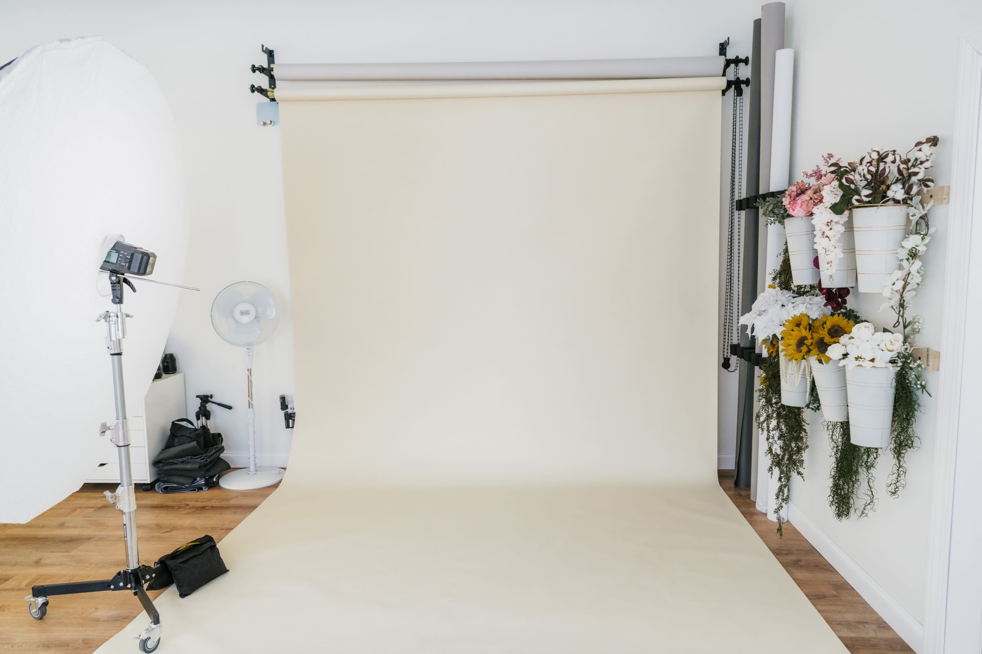 The image shows a photography studio with a beige backdrop, a light source on a stand, and decorative flower arrangements mounted on the wall.