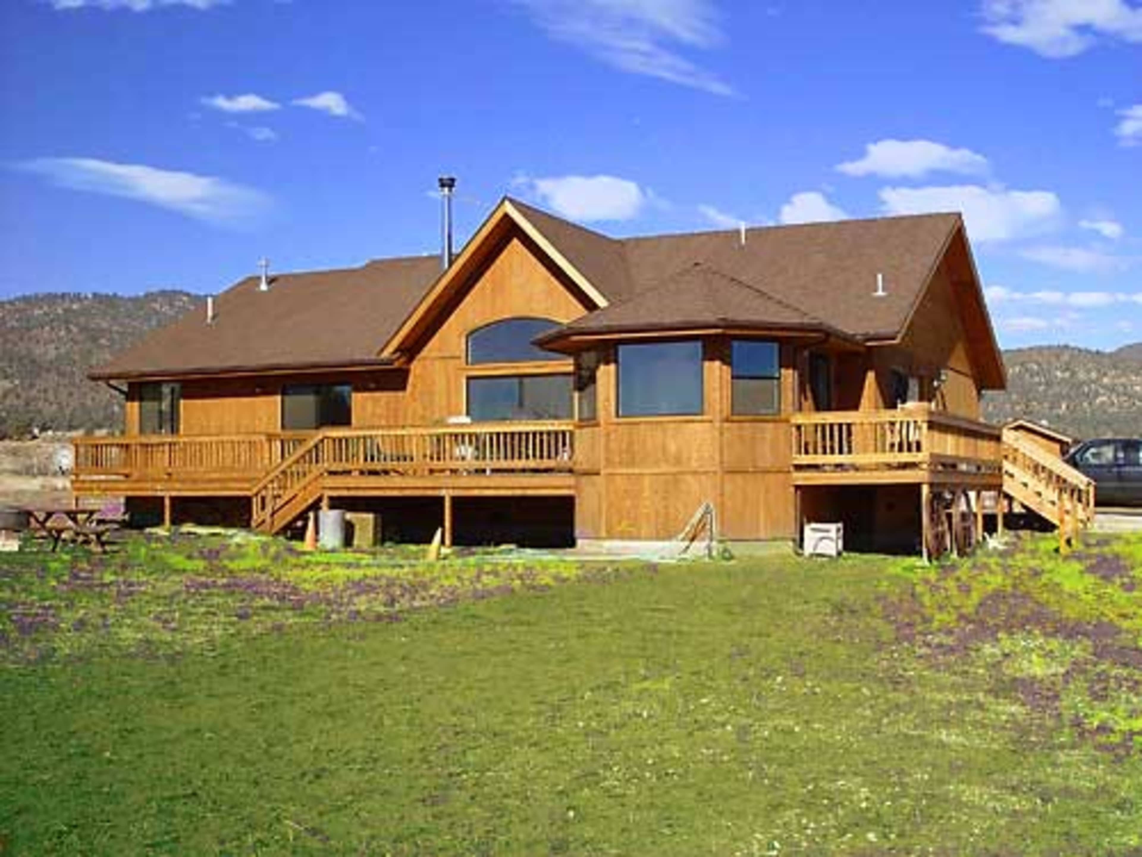 A large wooden house with multiple gabled roofs and a spacious deck overlooks a grassy yard against a mountainous backdrop.