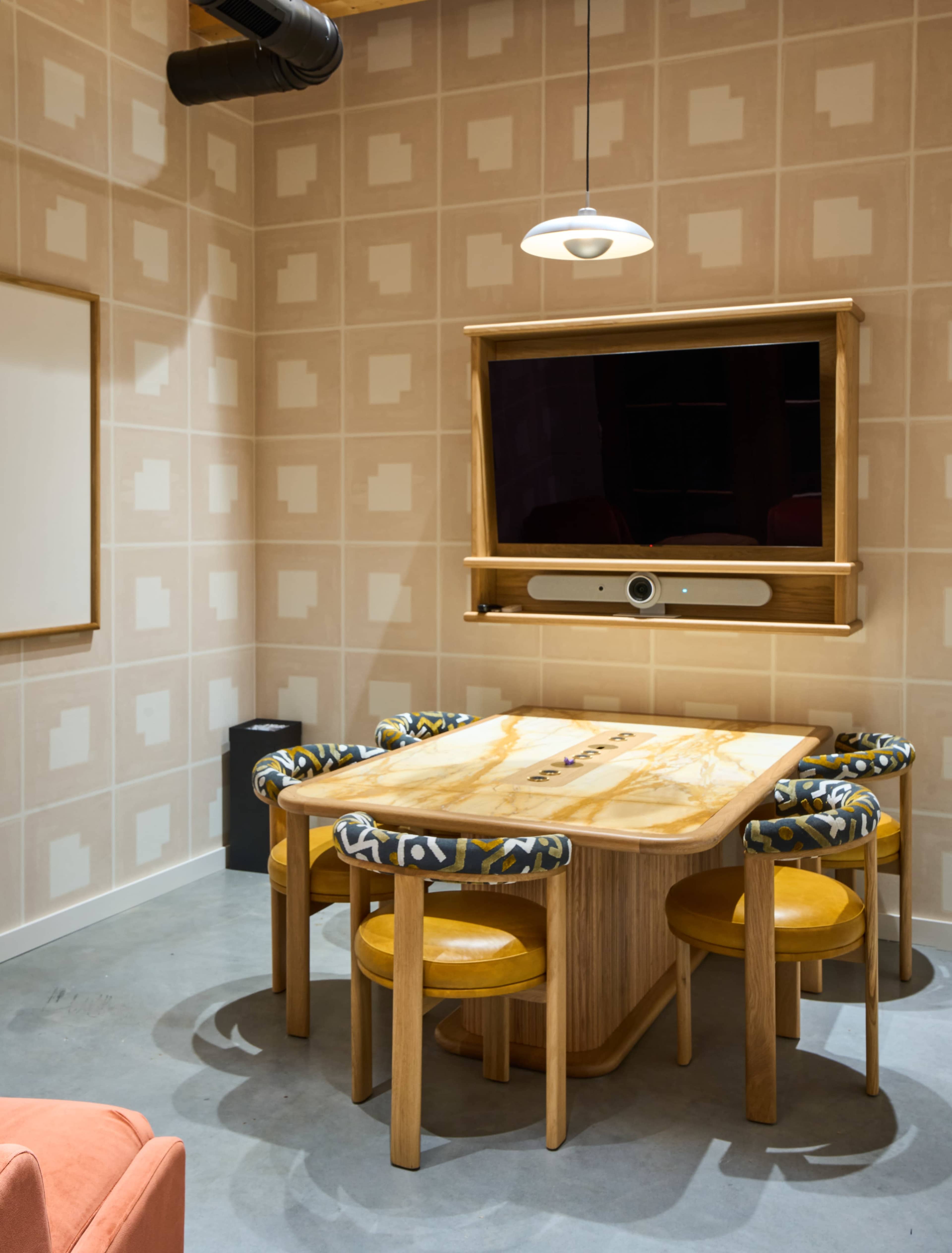 A small conference room features a wooden table with four chairs, a wall-mounted television, and patterned wallpaper.